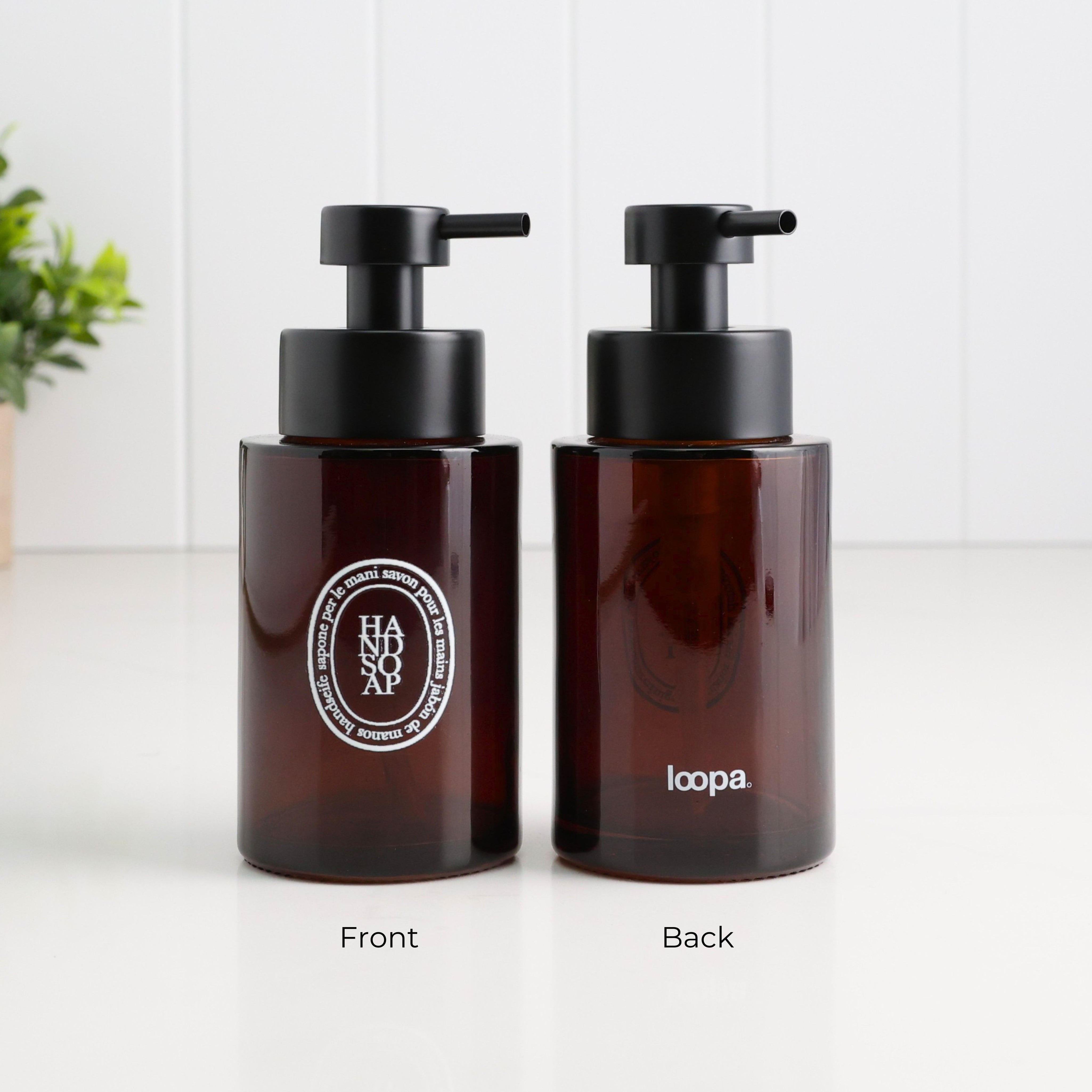 Front and back view of a 250ml amber glass refillable soap dispenser with white oval Hand Soap label, Loopa branding and satin black liquid pump, designed for bathroom, ensuite or kitchen use.