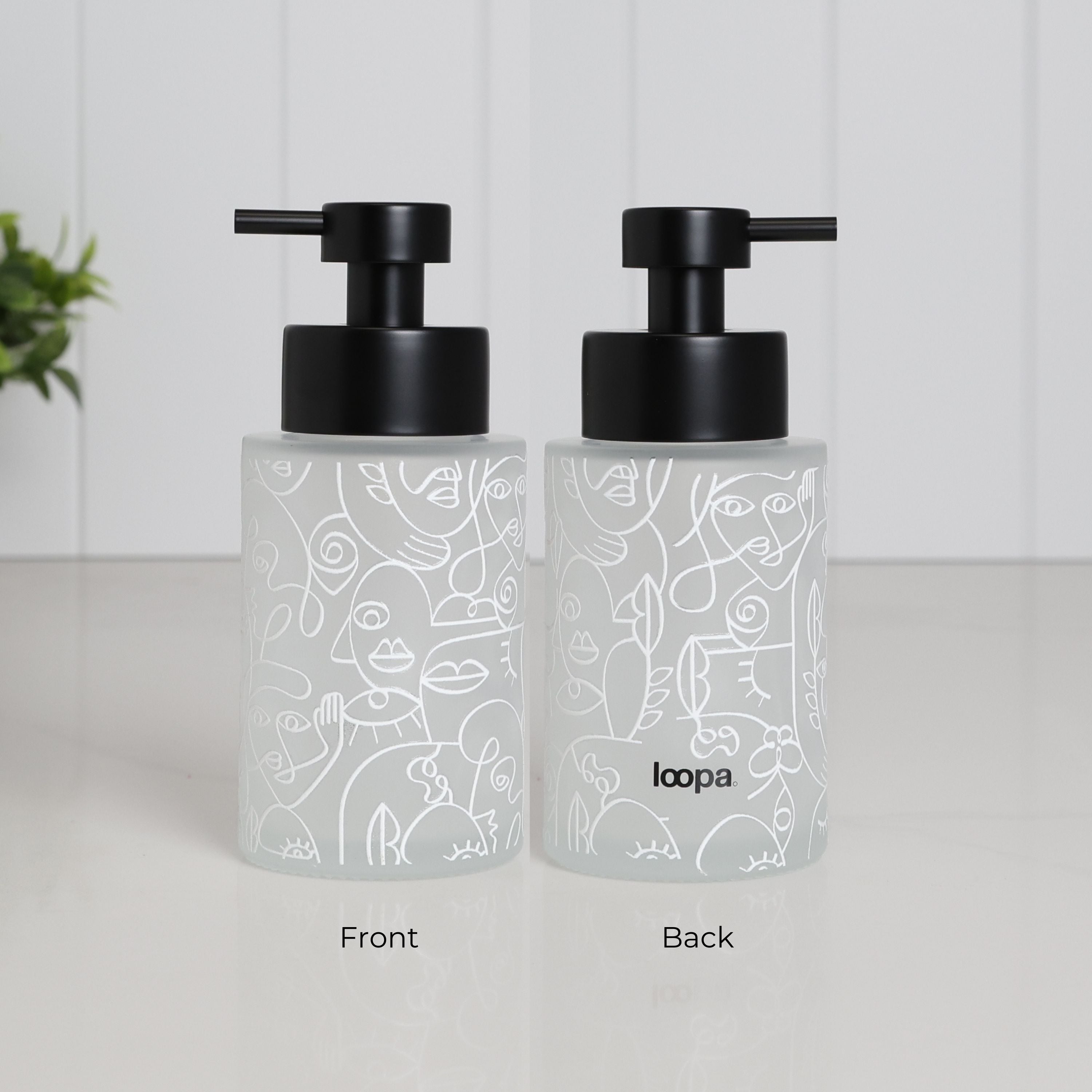 Front and back view of frosted glass soap dispenser with abstract face pattern wrap, 250ml refillable bathroom pump bottle featuring matte black stainless steel pump, modern reusable hand wash container with minimalist artistic design, eco friendly countertop accessory with subtle Loopa branding on reverse side