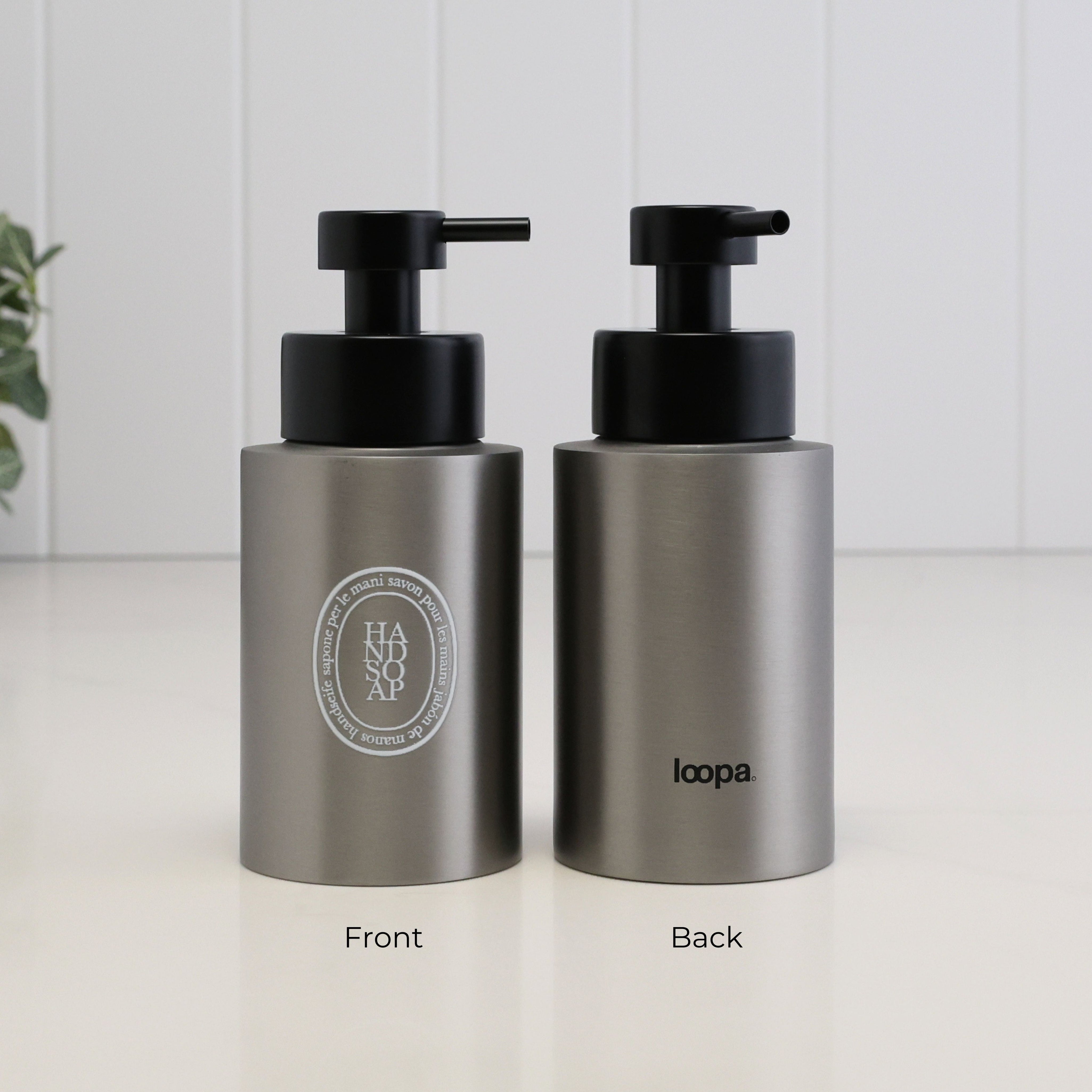 Two brushed stainless steel pump bottles shown side by side on a white surface against a white paneled wall. The left bottle shows the front with a circular logo reading HADSOAP, and the right bottle shows the back with the brand name loopa.