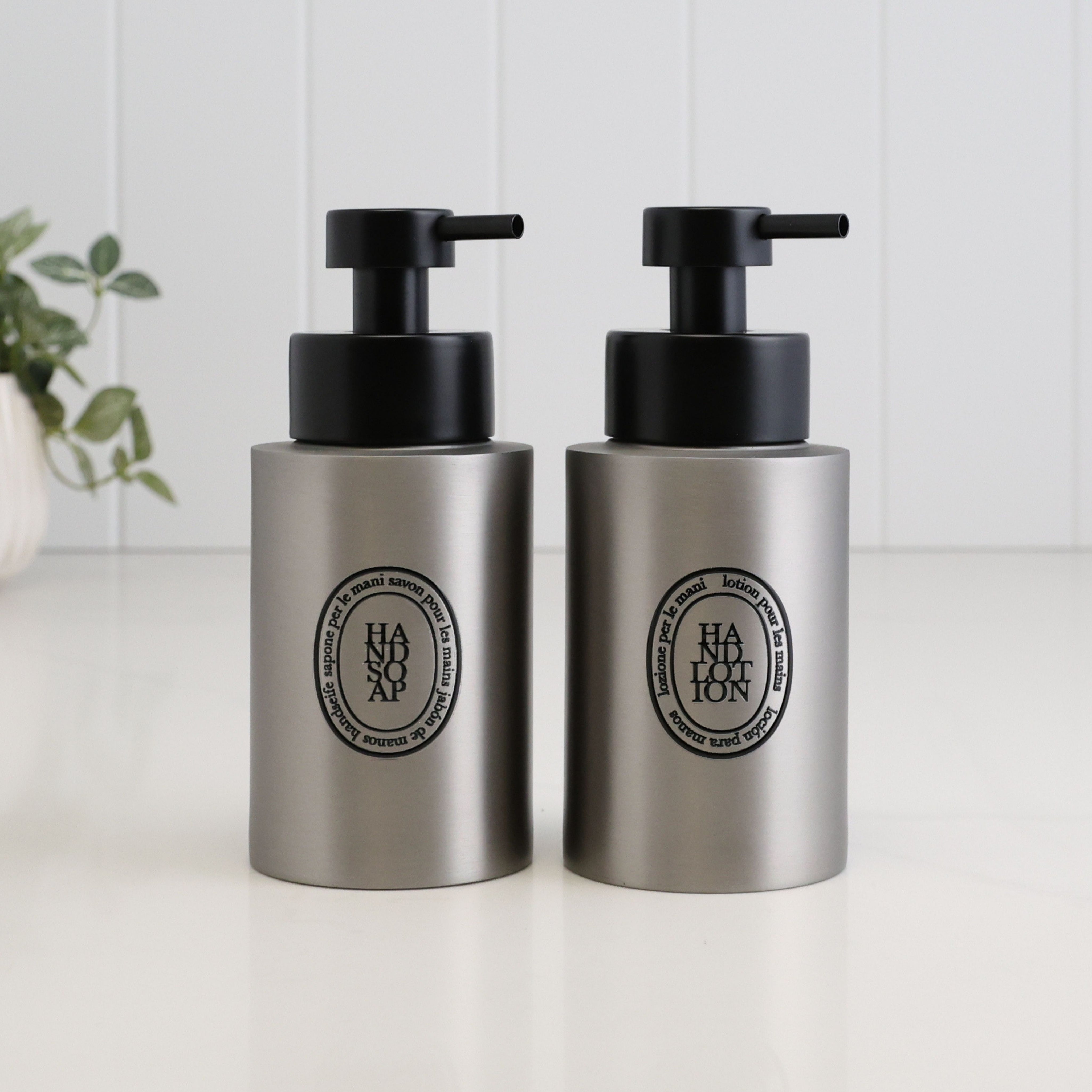 Pair of 250ml brushed 304 stainless steel hand soap and hand lotion dispensers with black oval labels and satin black liquid pumps, designed as a matching refillable bathroom or kitchen sink set.