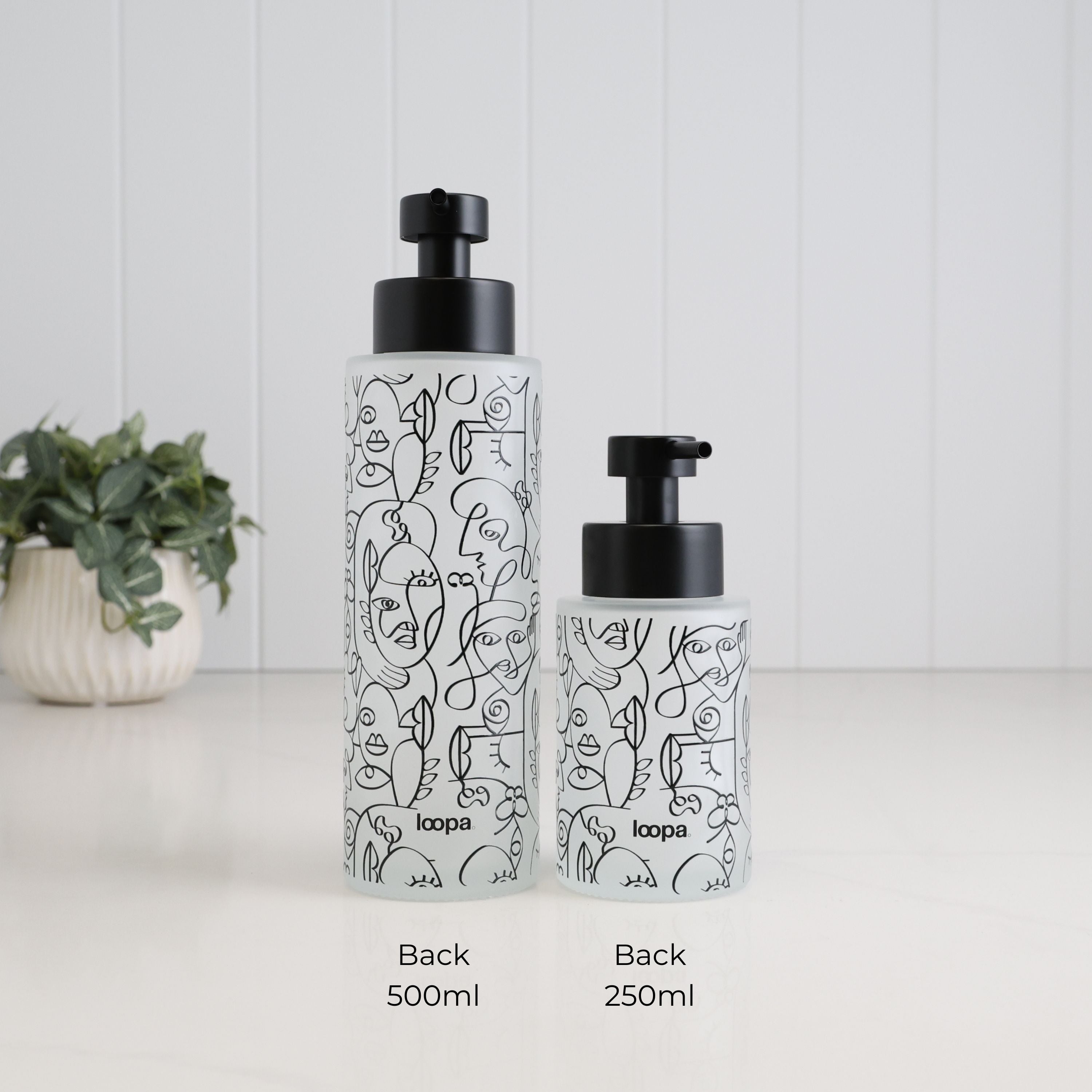 Two frosted glass pump bottles with black pump tops and black line-art face illustrations, labeled 500ml and 250ml, on a white surface against a white paneled background with a small potted plant at left.