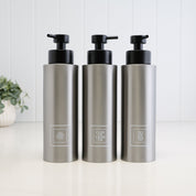 Set of three 500ml brushed 304 stainless steel shower dispensers for shampoo, conditioner and body wash, each with a white square label and satin black liquid pump.