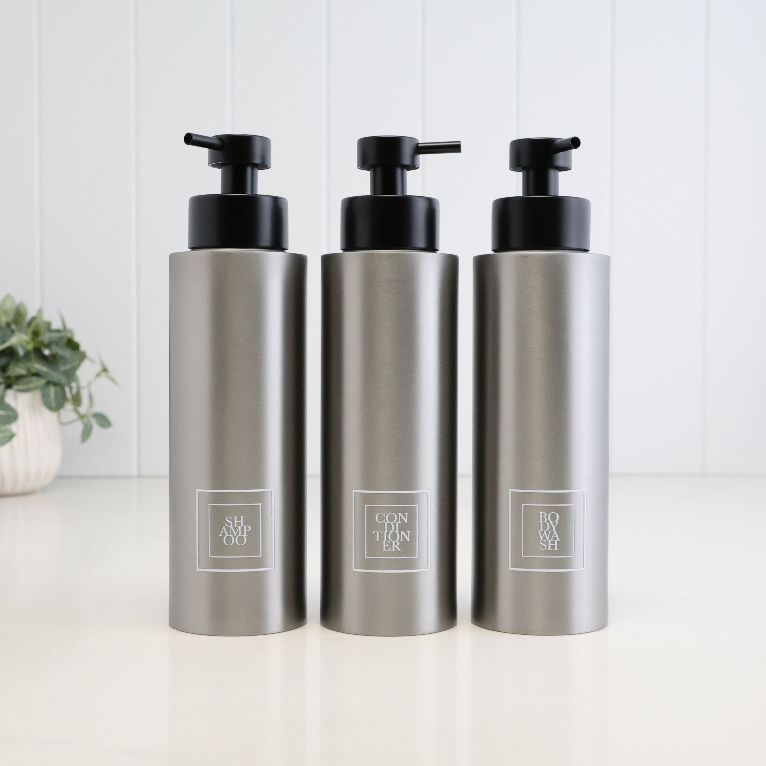Set of three 500ml brushed 304 stainless steel shower dispensers for shampoo, conditioner and body wash, each with a white square label and satin black liquid pump.