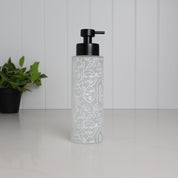 Tall frosted glass dispenser with abstract face line art wrap, 500ml refillable bathroom pump bottle with matte black stainless steel top, modern reusable shampoo or body wash container for minimalist shower space, eco friendly glass bottle with artistic pattern on clean white countertop