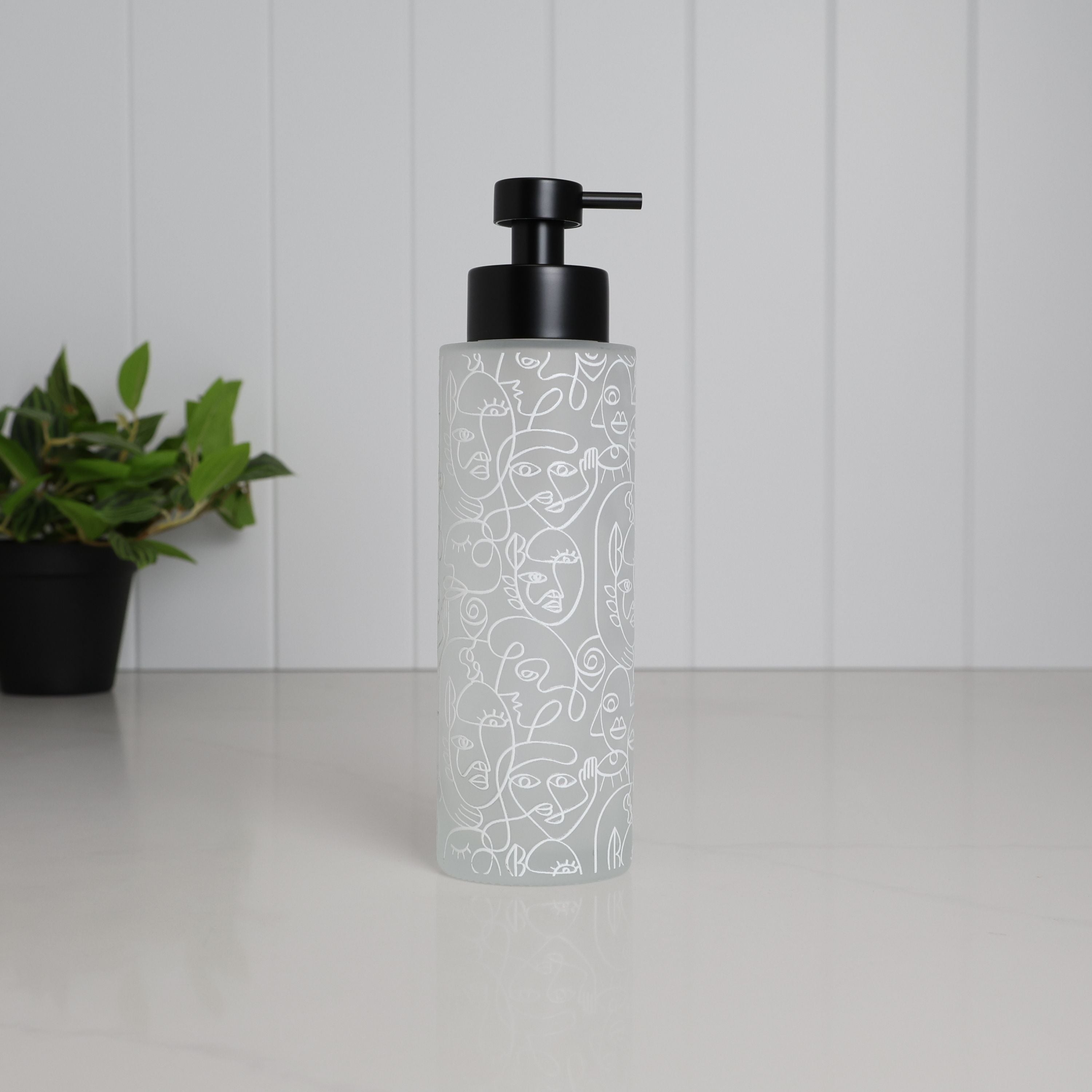 Tall frosted glass dispenser with abstract face line art wrap, 500ml refillable bathroom pump bottle with matte black stainless steel top, modern reusable shampoo or body wash container for minimalist shower space, eco friendly glass bottle with artistic pattern on clean white countertop