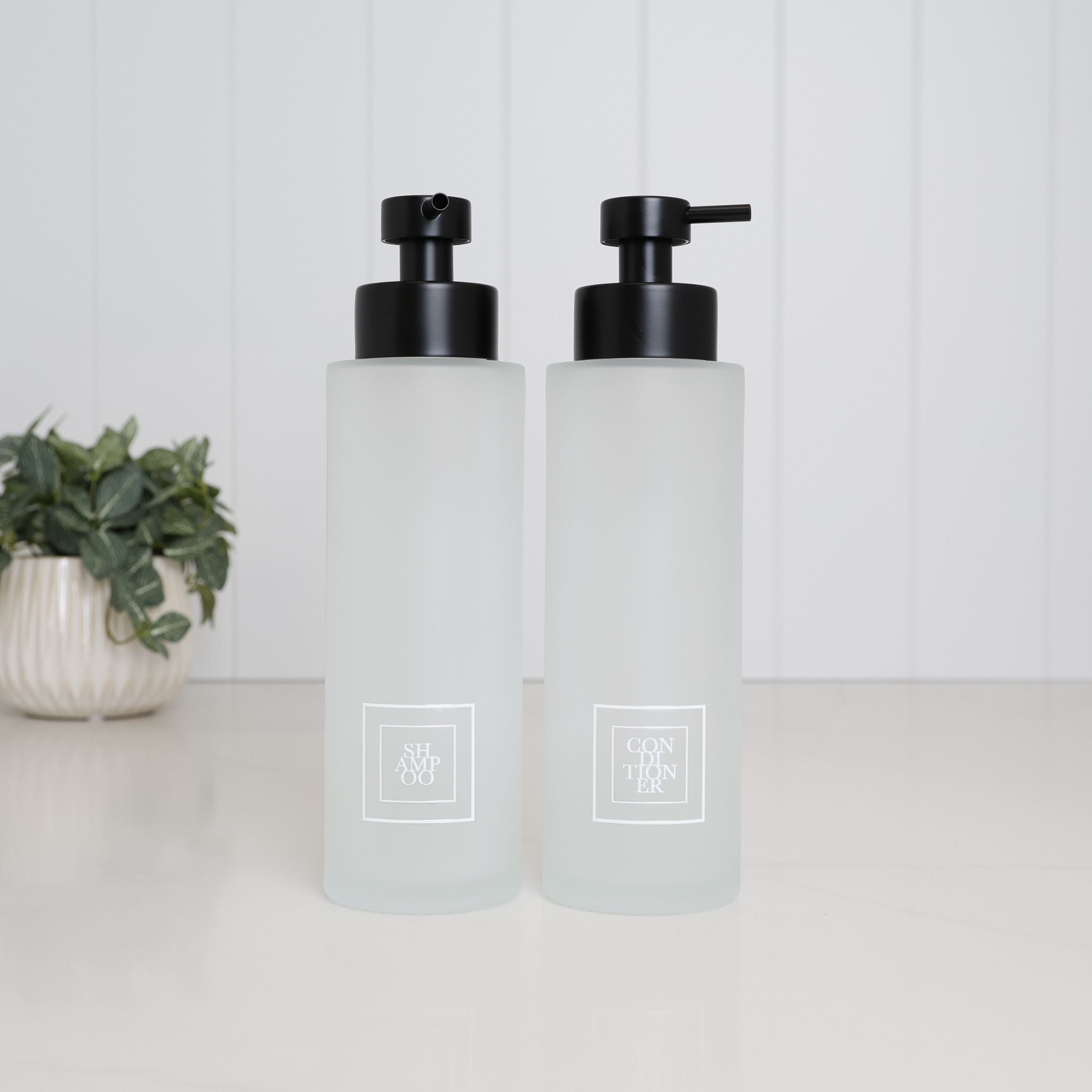 Set of two 500ml frosted glass soap dispensers with white Shampoo and Conditioner labels and satin black pumps, designed as matching refillable shampoo and conditioner bottles for a modern bathroom or shower.