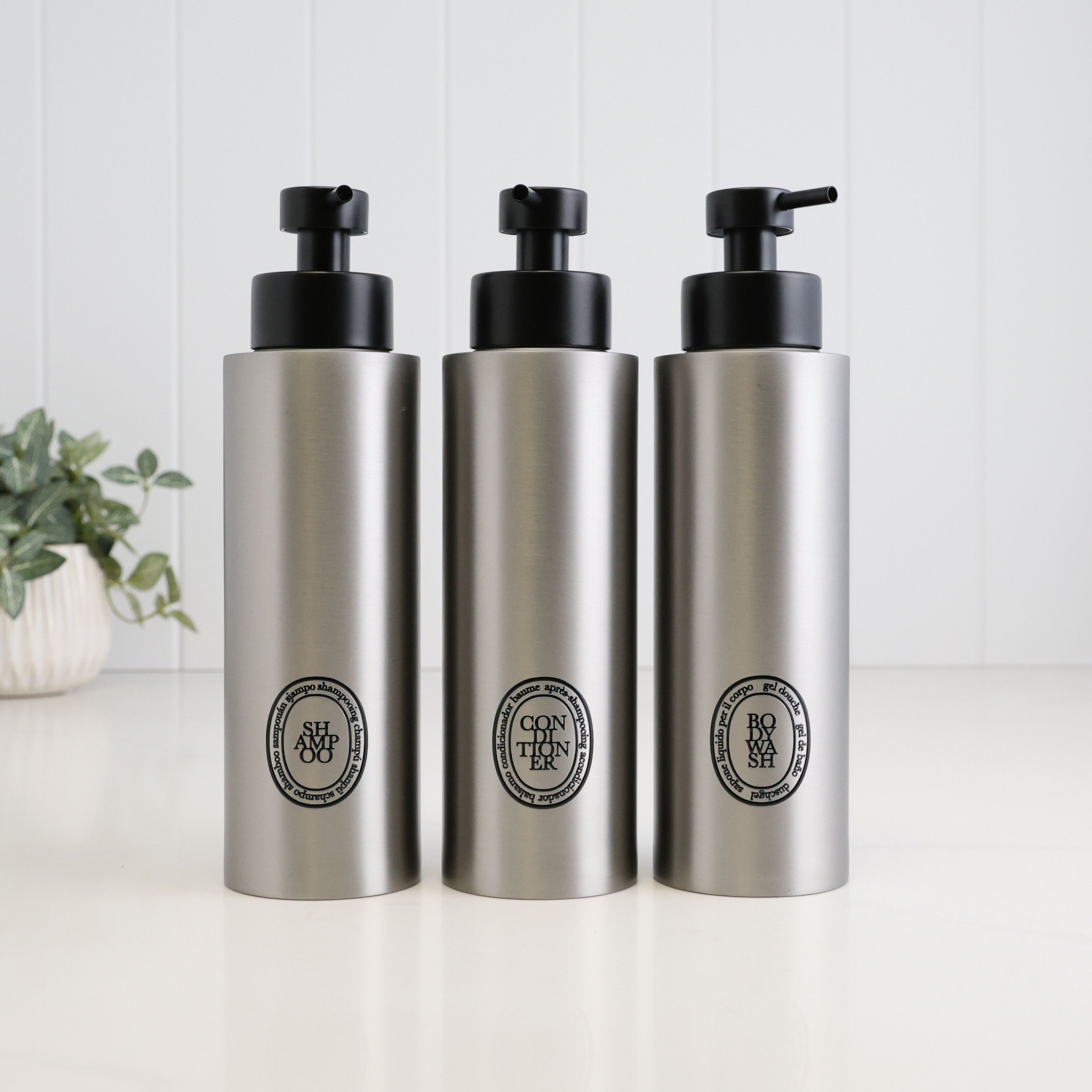 Set of three 500ml brushed 304 stainless steel refillable dispensers for shampoo, conditioner and body wash, with black oval labels and satin black liquid pumps, designed for an organised bathroom or shower.