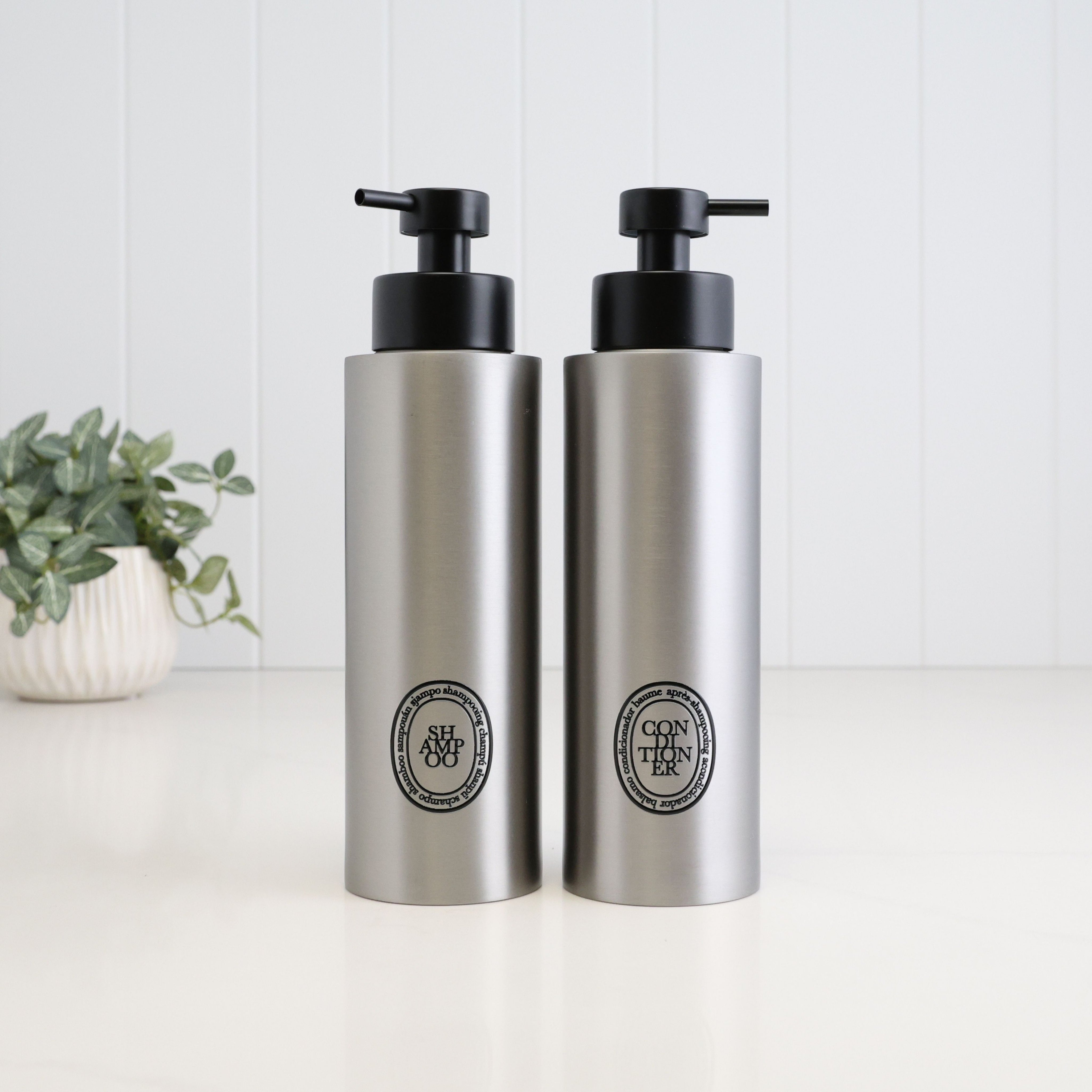 Pair of 500ml brushed 304 stainless steel refillable shampoo and conditioner dispensers with black oval labels and satin black liquid pumps, designed for coordinated bathroom and shower storage.