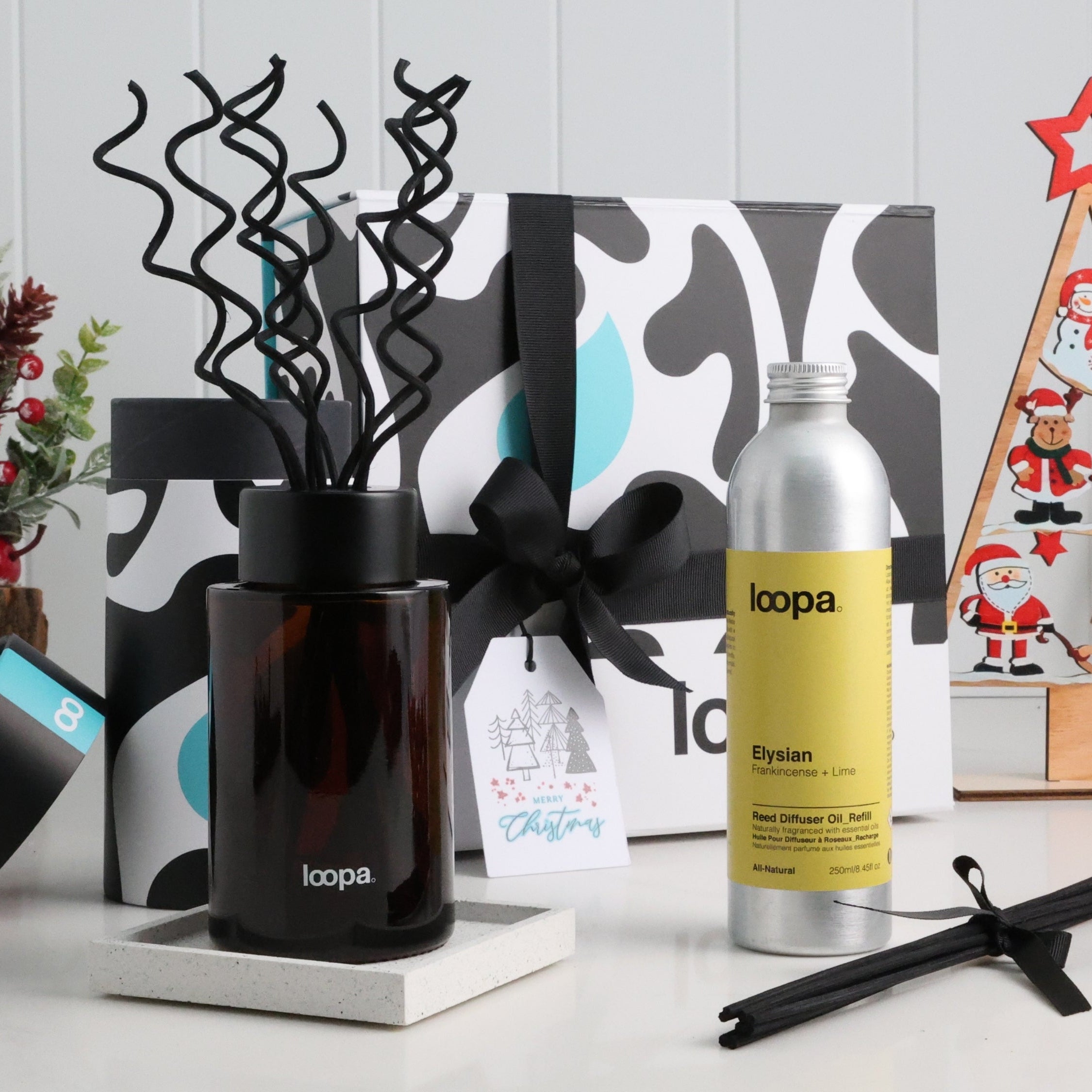 Amber glass reed diffuser with curled black reeds displayed beside Elysian refill and a festive Loopa gift box. Styled with Christmas décor, ideal as a luxury holiday home fragrance present for cosy winter gifting.