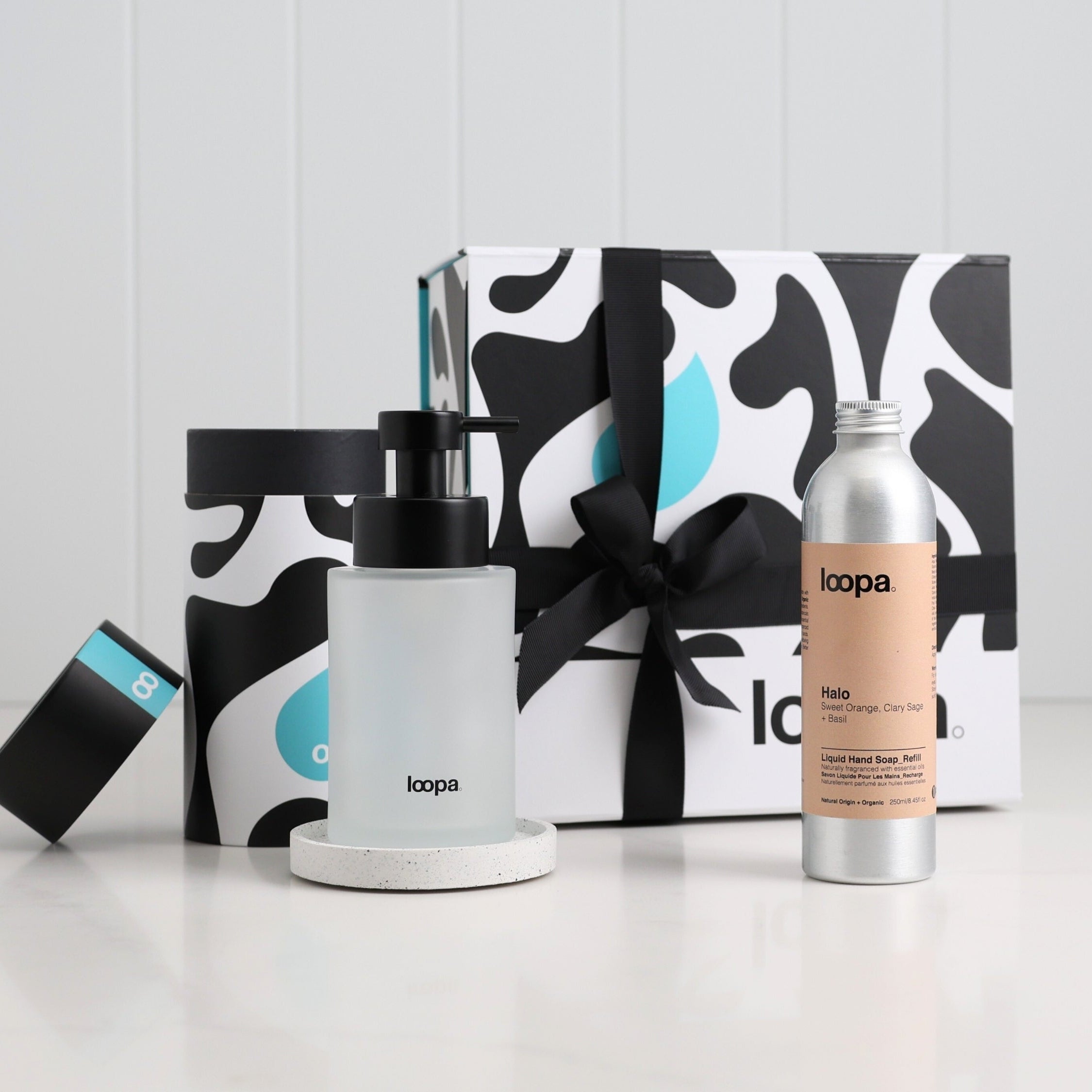 Frosted glass soap dispenser on a terrazzo-style base beside a Halo hand soap refill bottle, styled with Loopa’s black-and-white gift box and ribbon for a clean, modern premium gifting look.