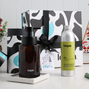 Amber glass soap dispenser and refill bottle in festive gift box with black ribbon and Christmas tag on white surface.