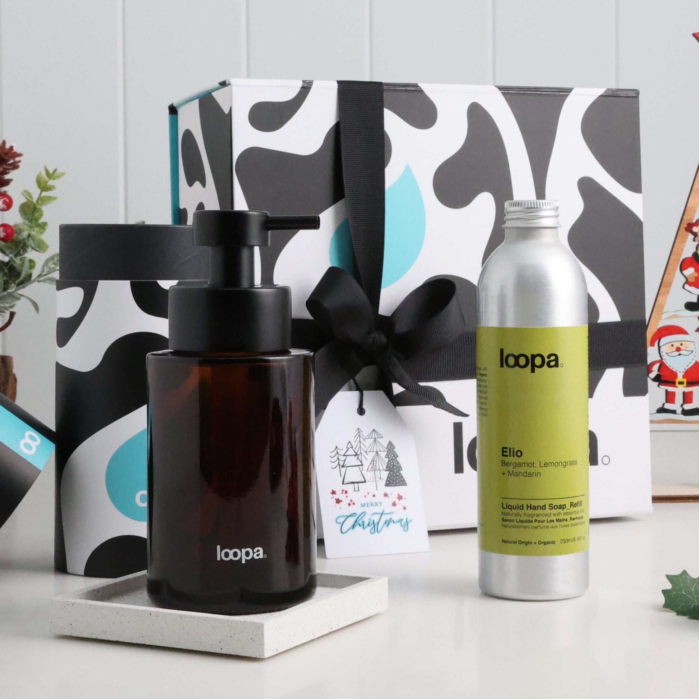 Amber glass soap dispenser and refill bottle in festive gift box with black ribbon and Christmas tag on white surface.