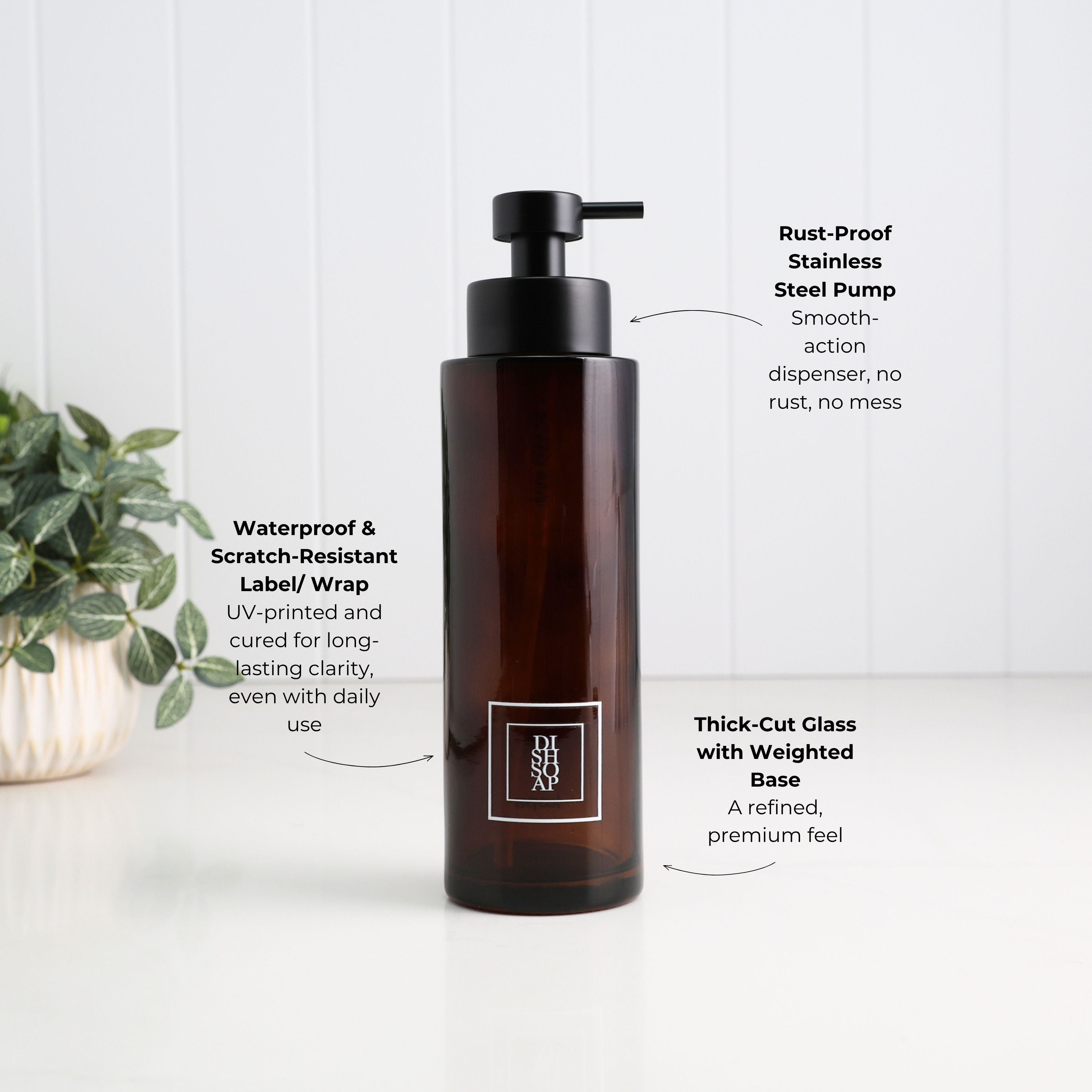 An amber brown pump bottle with a black dispenser, shown on a white surface against a white paneled wall. Text callouts describe product features, and a small green plant is visible on the left.