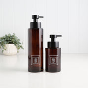 Two amber glass pump bottles with black pumps and white square labels reading HANXO, standing on a white surface against a white paneled wall, with a small potted plant in the background.
