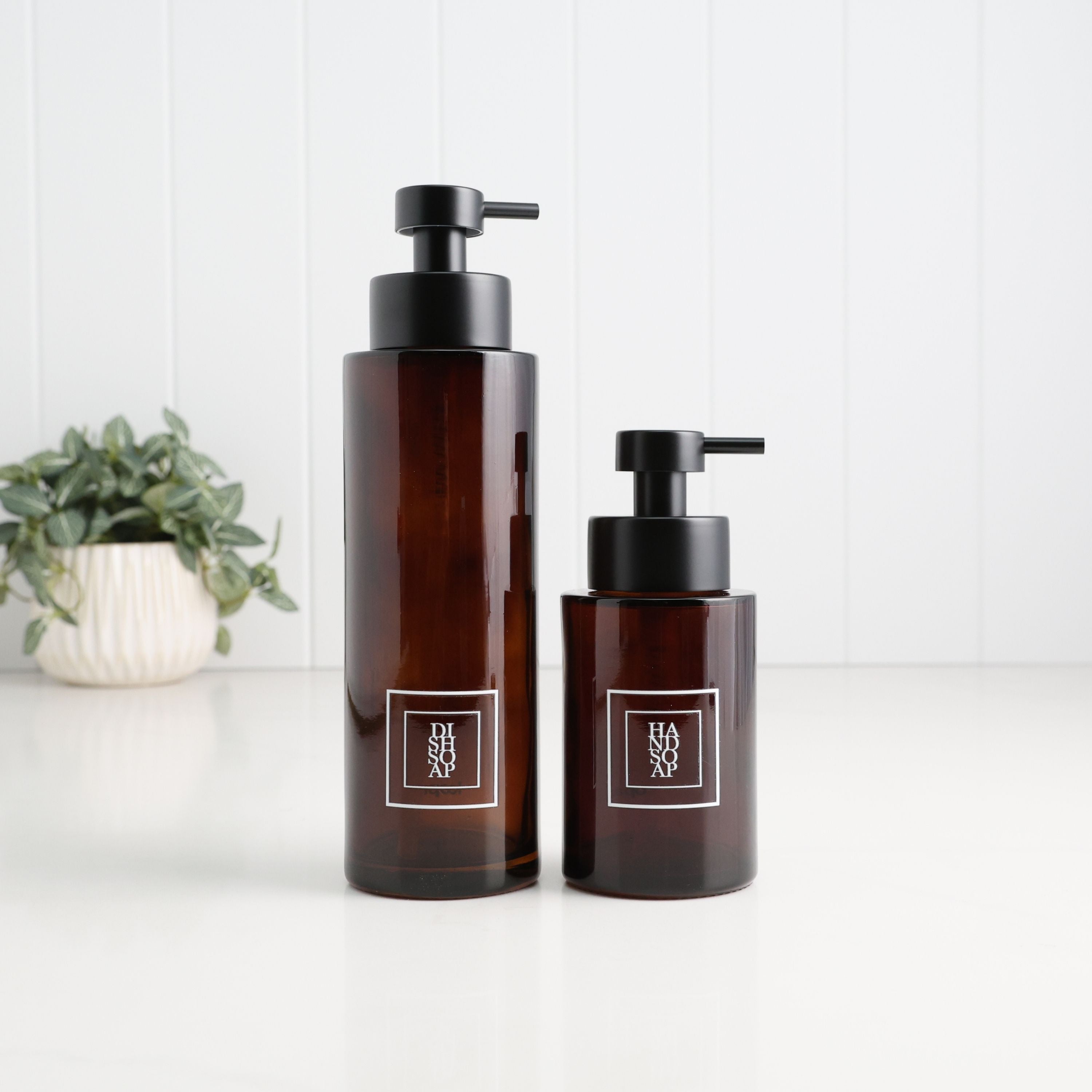 Two amber glass pump bottles with black pumps and white square labels reading HANXO, standing on a white surface against a white paneled wall, with a small potted plant in the background.