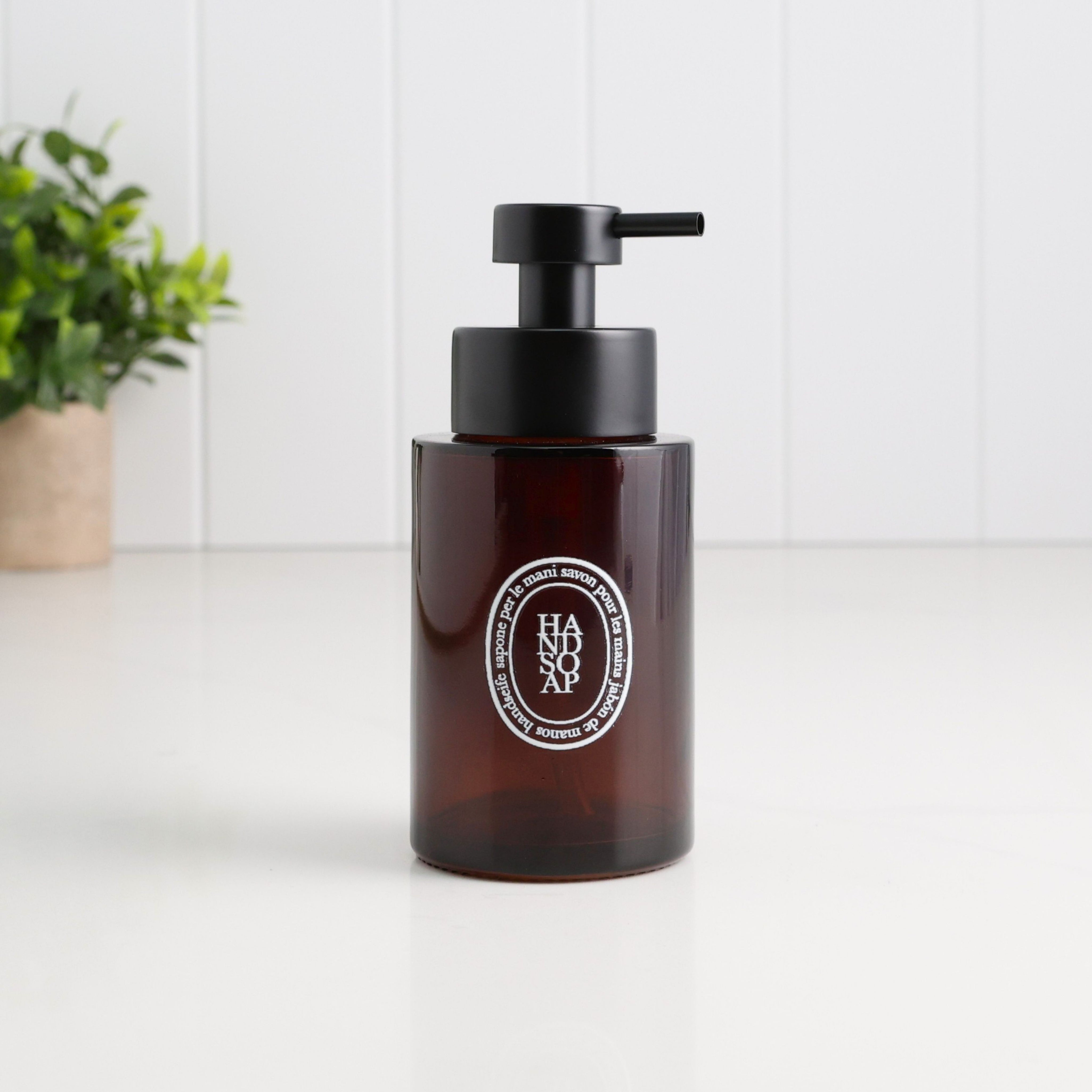 Amber glass 250ml refillable hand soap dispenser with white oval Hand Soap label and satin black stainless steel liquid pump, styled on a clean white surface for a modern bathroom, cloakroom or kitchen.