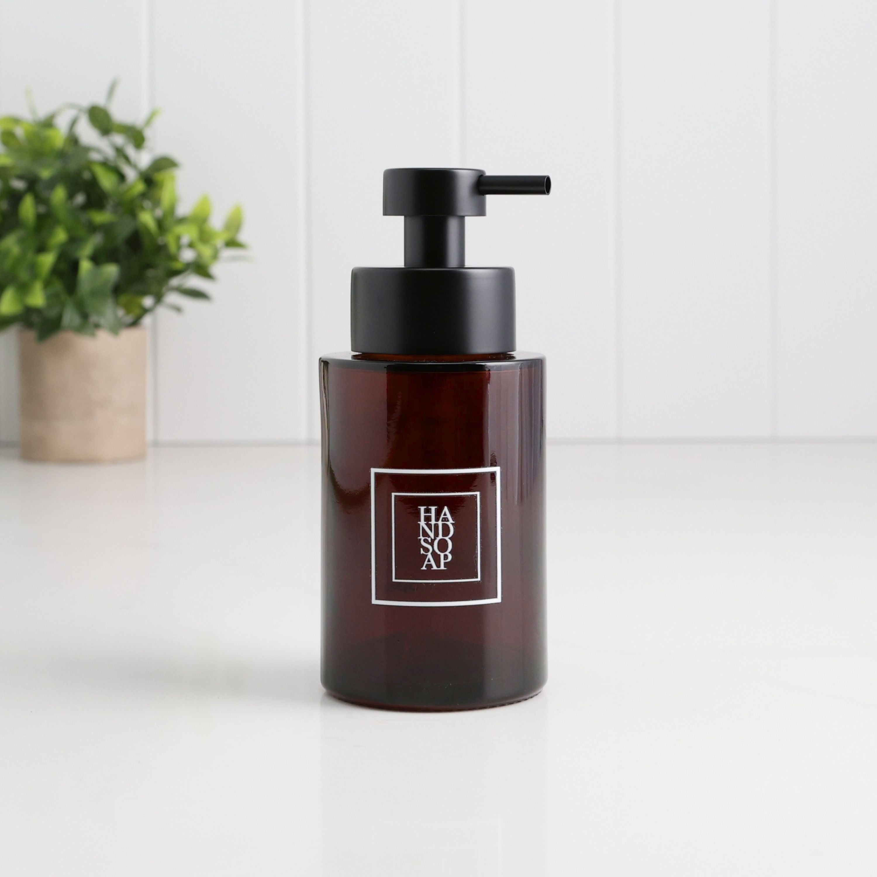 An amber glass pump bottle with a black dispenser and a white square logo label reading 'HAIR SOAP', set on a white surface with a blurred green plant in the background.