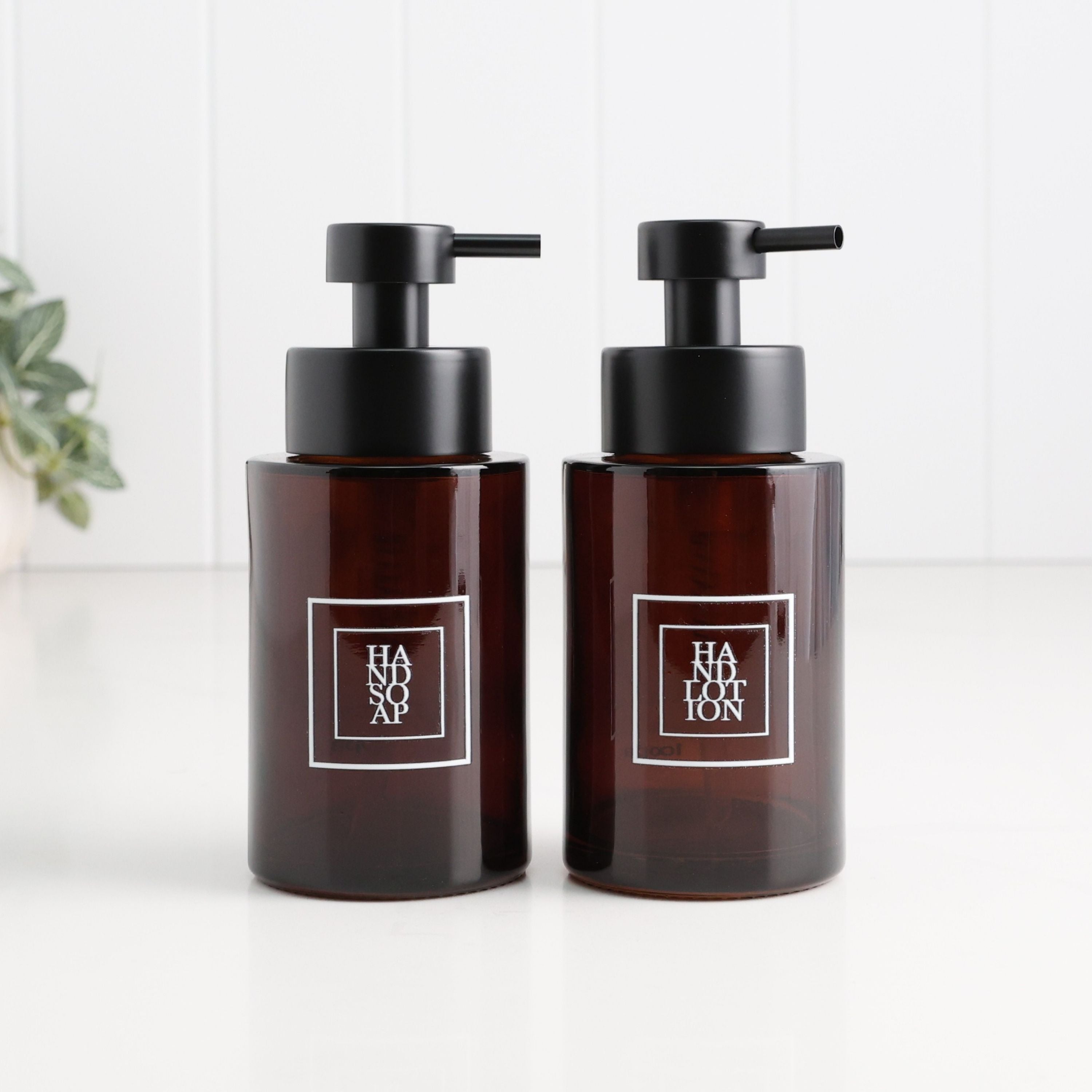 Two amber glass pump bottles with black dispensers and white square labels reading HANSON, standing on a white surface against a white paneled background.