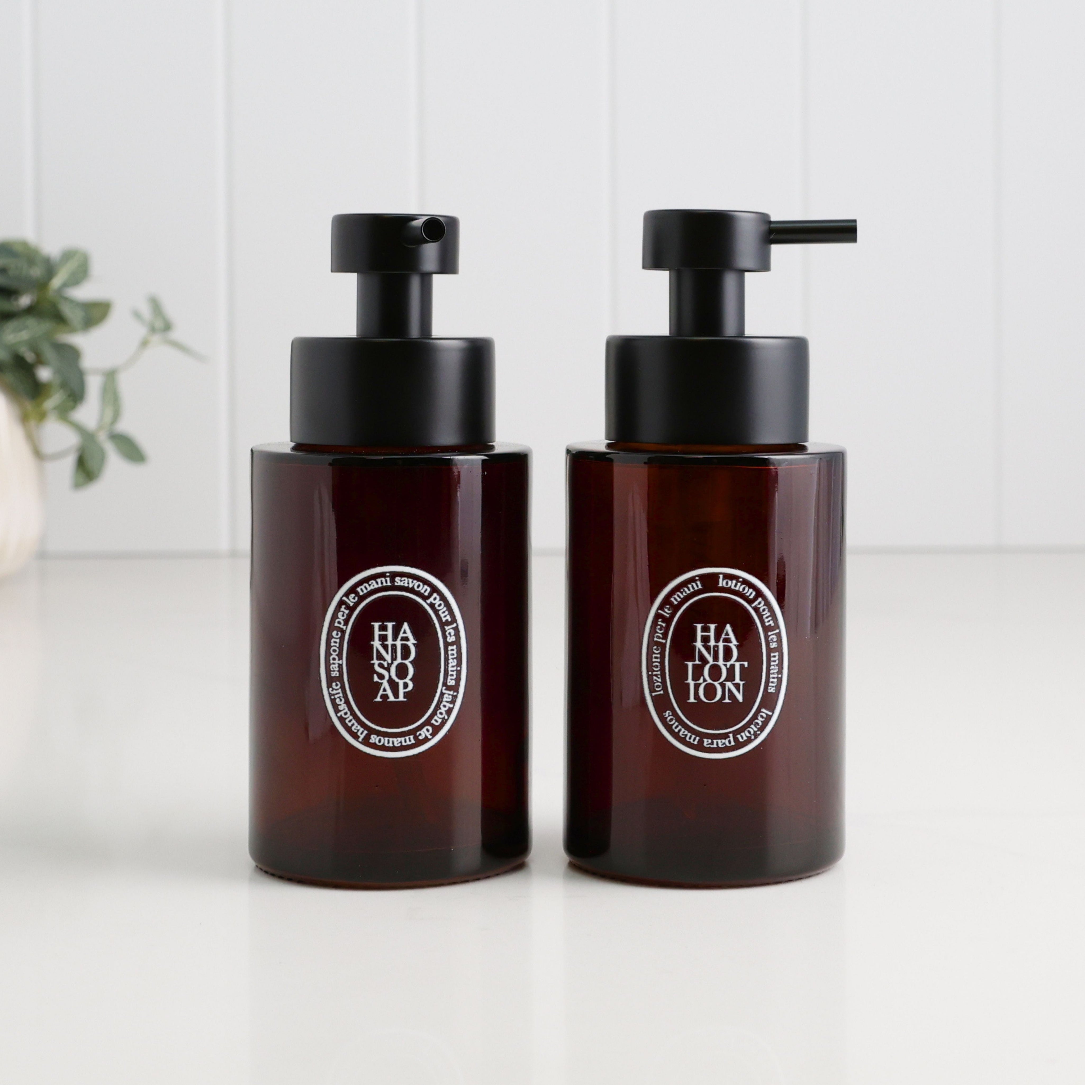 Pair of 250ml amber glass refillable dispensers for hand soap and hand lotion, featuring white oval labels and satin black liquid pumps for a coordinated bathroom or kitchen sink set.