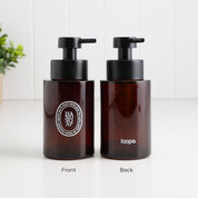 Front and back views of a 250ml amber glass refillable soap dispenser, featuring a white oval Hand Soap label, Loopa branding and satin black stainless steel liquid pump.