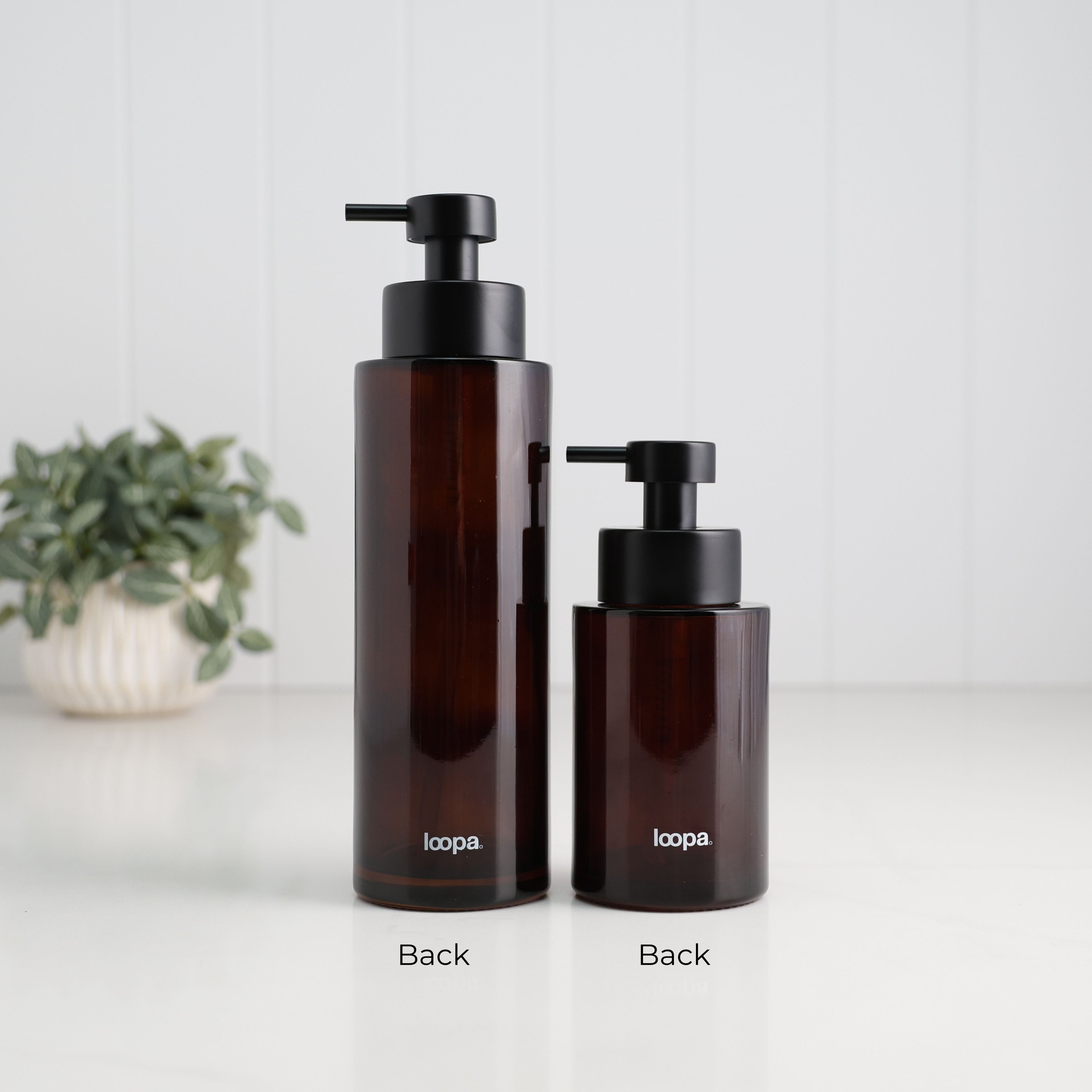 Back view of 500ml and 250ml amber glass refillable soap dispensers with Loopa branding and satin black stainless steel liquid pumps, showing the two bottle sizes side by side.
