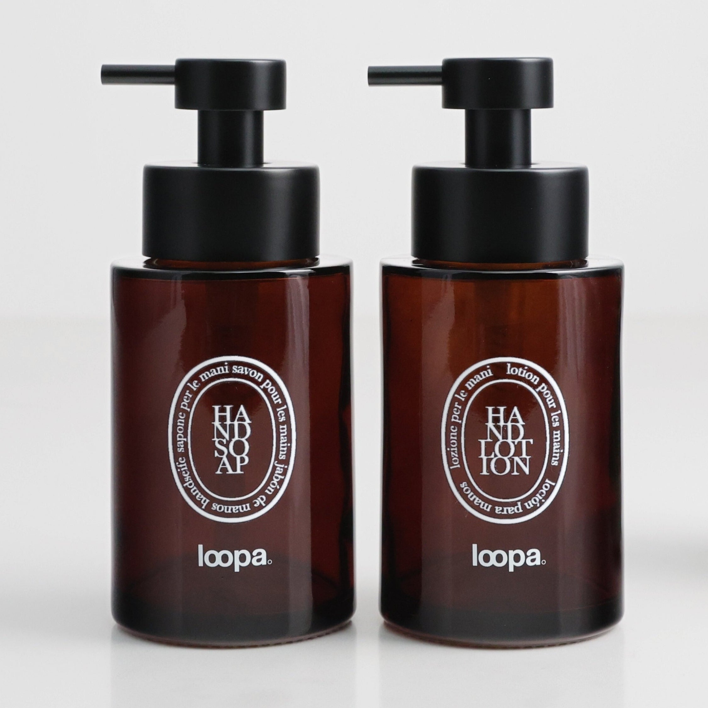 Two amber glass round bottle dispensers with white oval labels, presented as a coordinated 250ml refillable set for bathrooms, cloakrooms, or kitchen use