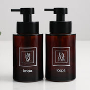 Two amber glass round bottle dispensers with white square labels, shown together as matching 250ml refillable options for soap or lotion