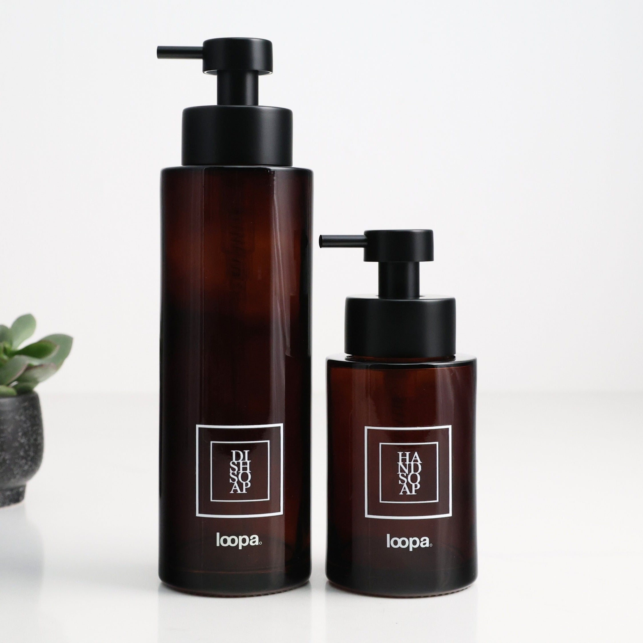 Two amber glass round bottle dispensers with white square labels, shown together in 500ml and 250ml refillable sizes for soap or lotion