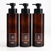 Three amber glass round bottle dispensers with white square labels, shown together as matching 500ml refillable options for soap or lotion