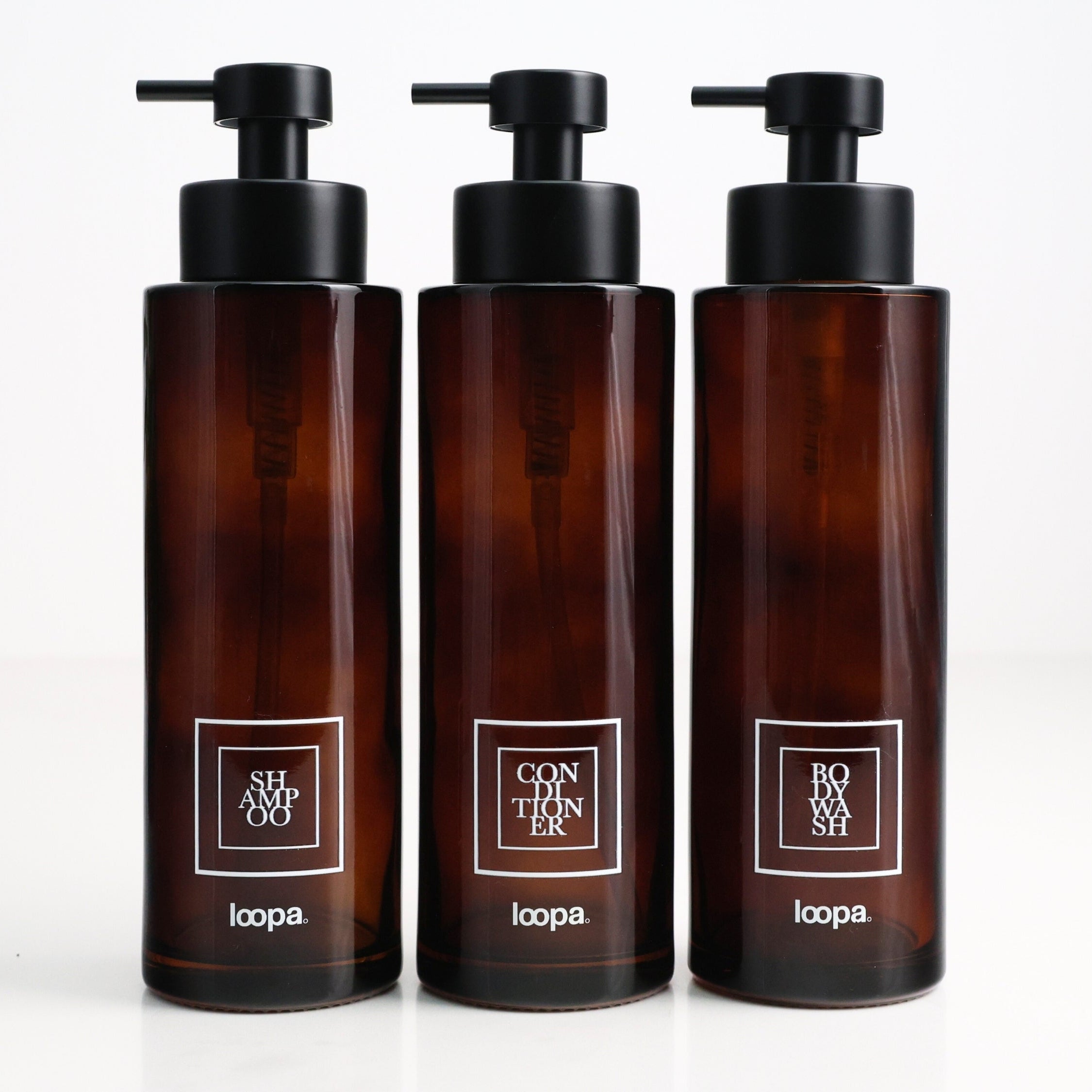 Three amber glass round bottle dispensers with white square labels, shown together as matching 500ml refillable options for soap or lotion