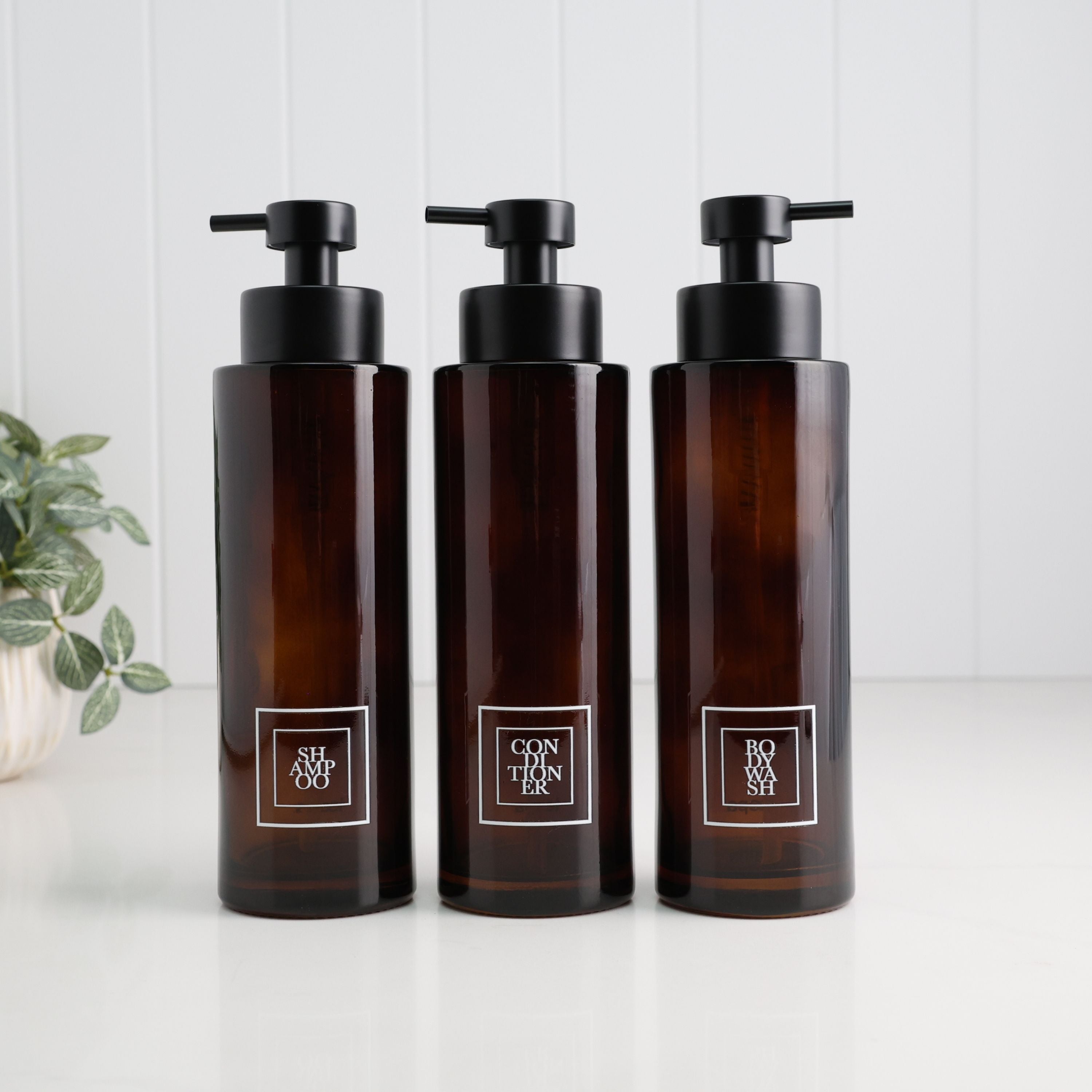 Three amber glass pump bottles with black dispensers and white labels, arranged side by side on a white surface against a white paneled background.