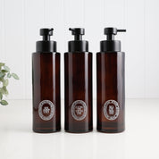 Three amber glass pump bottles with black dispensers and oval white labels, arranged side by side on a white surface against a white paneled background.