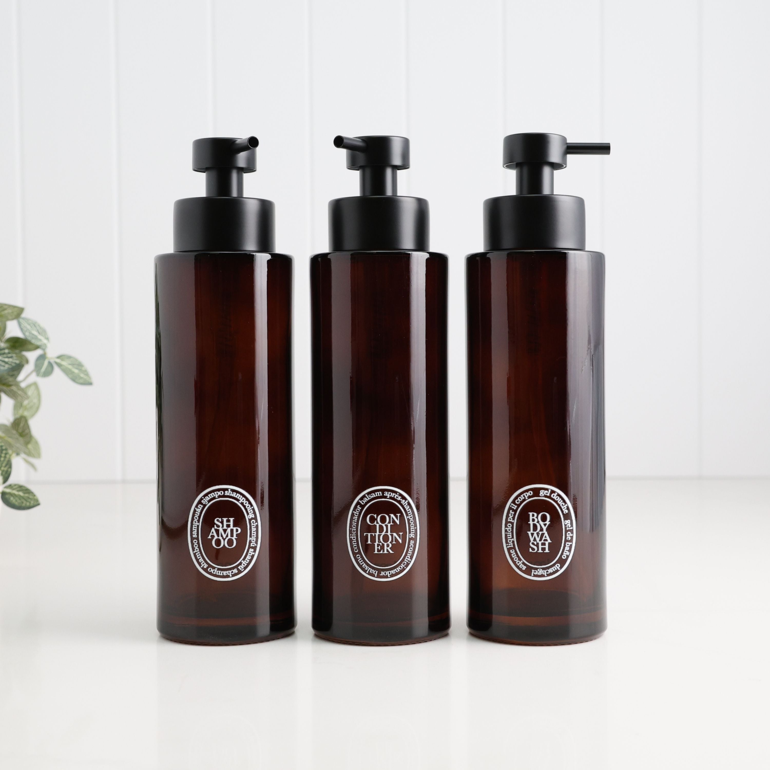 Three amber glass pump bottles with black dispensers and oval white labels, arranged side by side on a white surface against a white paneled background.