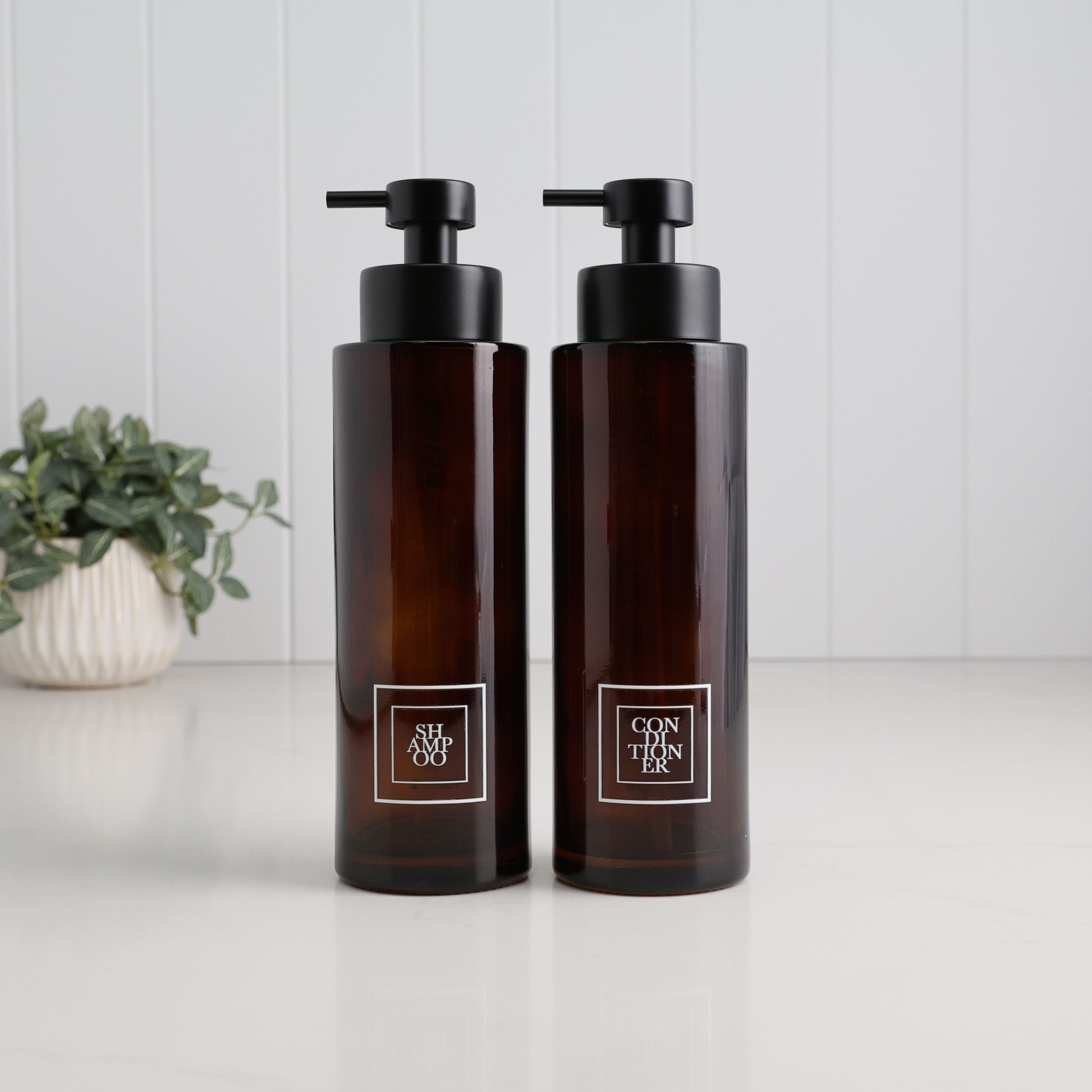 Two amber glass pump bottles with black dispensers standing side by side on a white surface against a white paneled wall. Each bottle has a square white label with black text; the left label reads 'THE ALOHO' and the right label reads 'CON TIAN ER'.