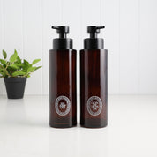 Two amber glass pump bottles with black dispensers and circular silver Diptyque labels, standing on a white surface against a white paneled background with a small green plant in the rear left.