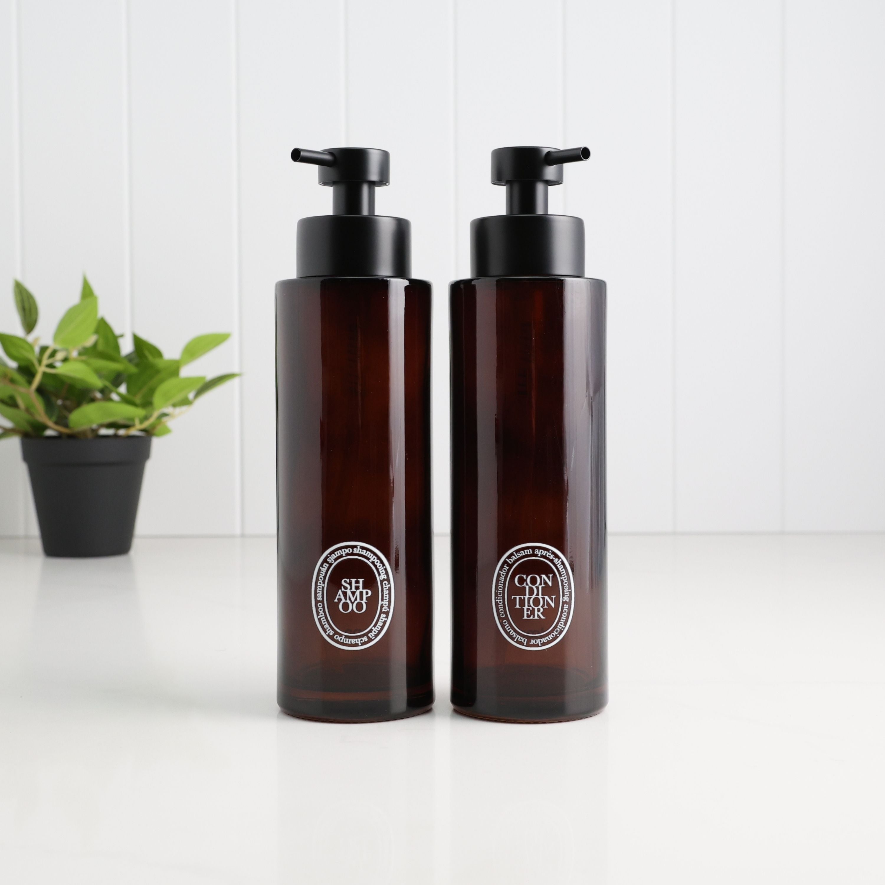 Two amber glass pump bottles with black dispensers and circular silver Diptyque labels, standing on a white surface against a white paneled background with a small green plant in the rear left.