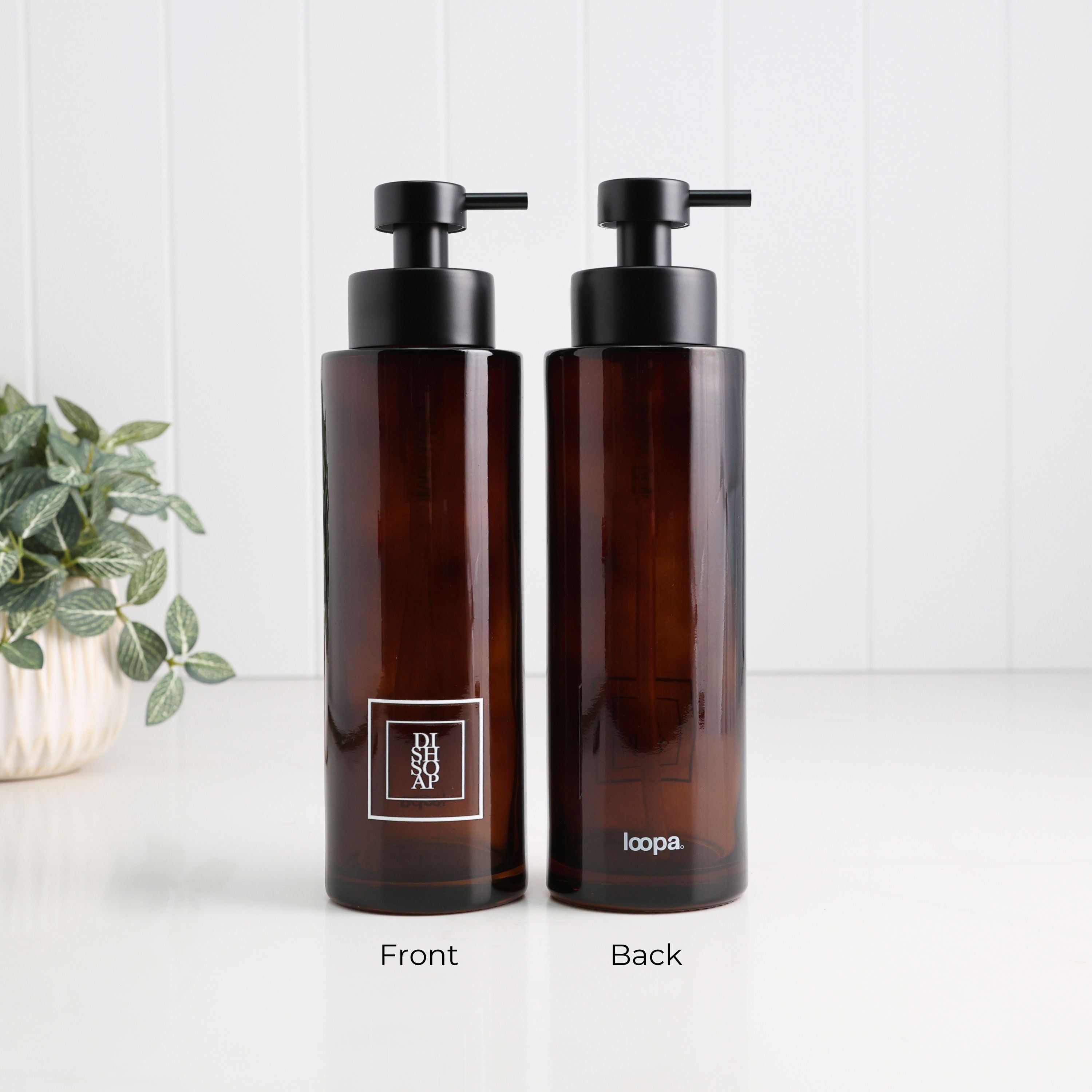 Two amber glass pump bottles shown side by side on a white surface with a white paneled wall behind. The left bottle is labeled Front and shows the front of a square white logo patch; the right bottle is labeled Back and shows the back of the same bottle.