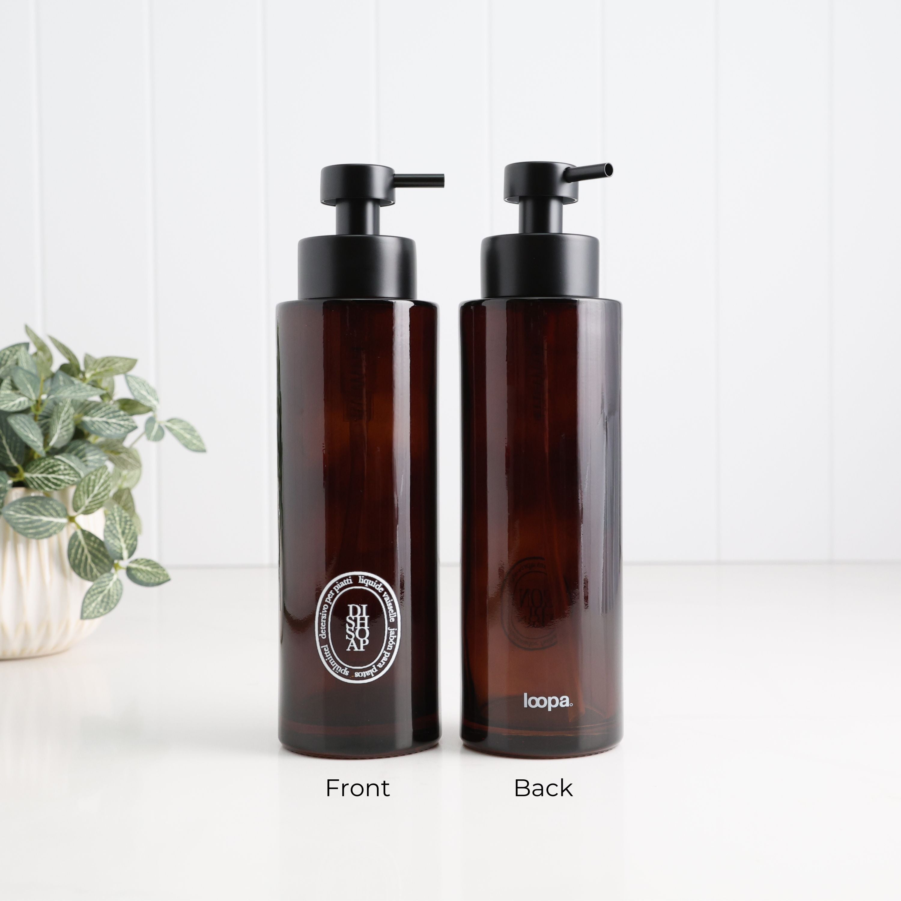 Two amber glass pump bottles shown side by side on a white surface against a white paneled wall, labeled Front and Back. A small potted plant sits on the left.
