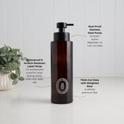 A dark amber glass pump bottle with a black dispenser, shown on a white surface against a white paneled wall. Text callouts describe its features: rust-proof stainless steel pump, waterproof and scratch-resistant label/wrap, and thick-cut glass with a weighted base.