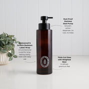 A dark amber glass pump bottle with a black dispenser, shown on a white surface against a white paneled wall. Text callouts describe its features: rust-proof stainless steel pump, waterproof and scratch-resistant label/wrap, and thick-cut glass with a weighted base.