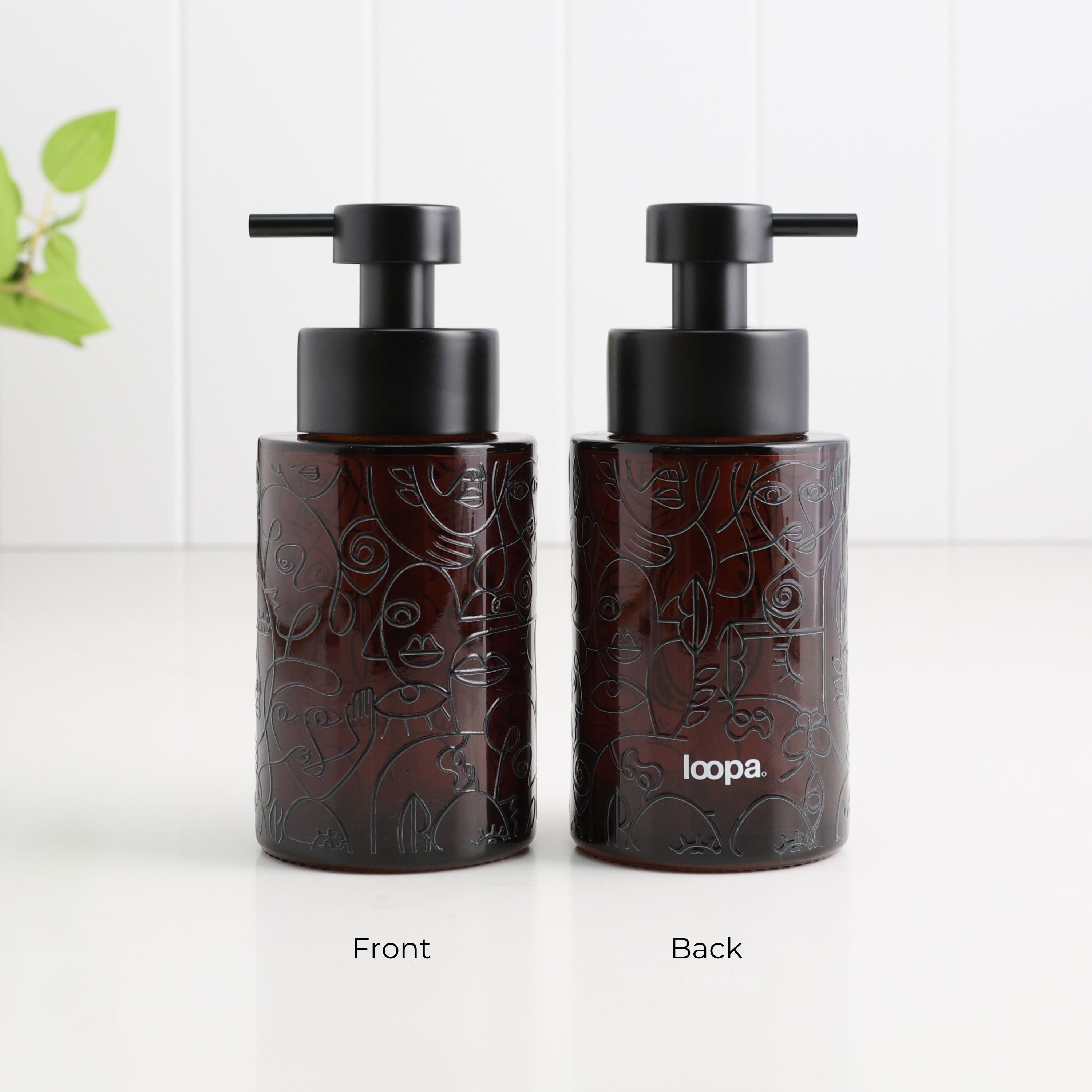Two dark amber glass pump bottles with black pumps, shown side by side on a white surface against a white paneled background. The left bottle is labeled Front and shows an embossed floral pattern, while the right bottle is labeled Back and shows the white 'loopa.