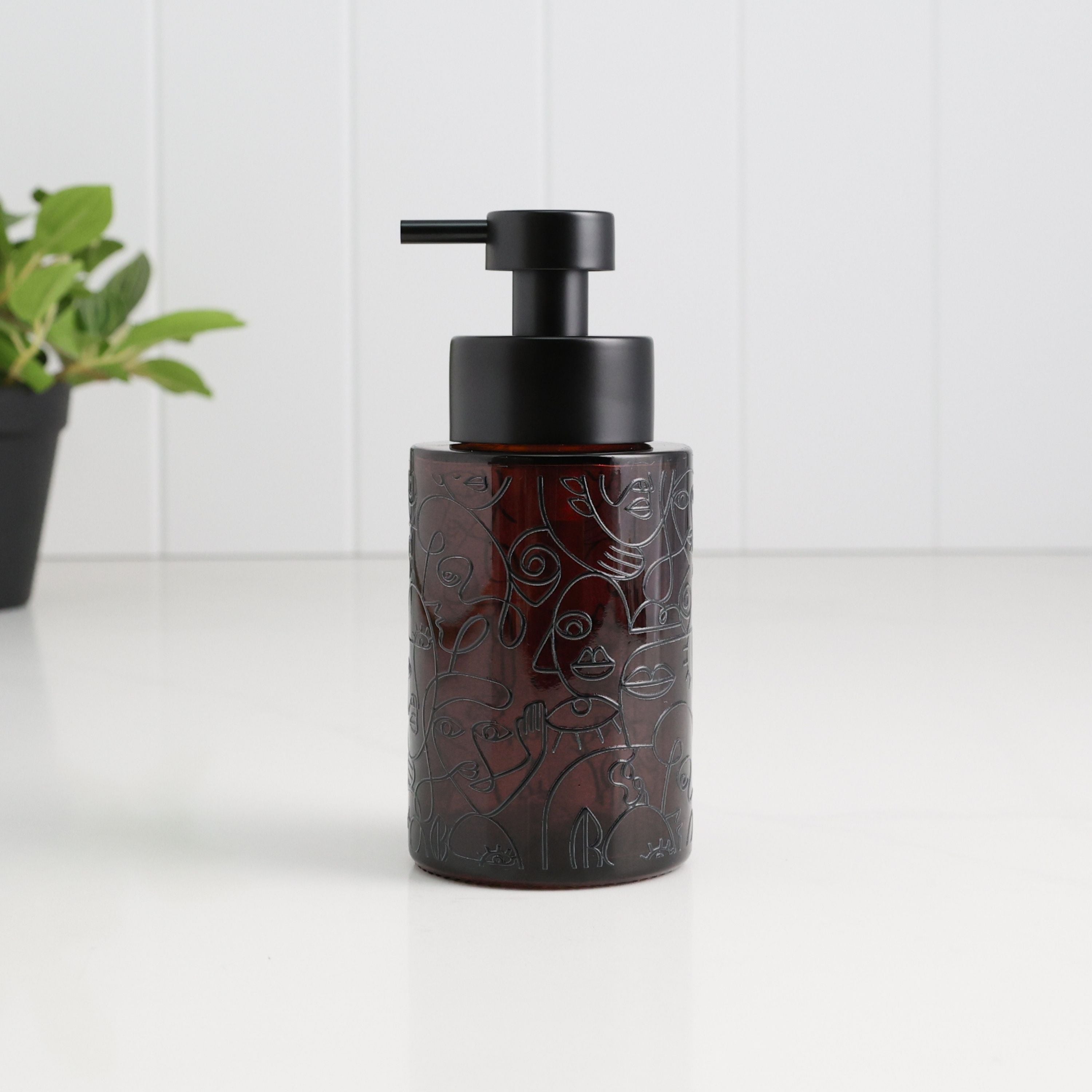 A dark amber glass soap dispenser with a black pump, featuring an embossed abstract face pattern, set on a white surface with a white paneled wall background and a small potted plant in the distance.