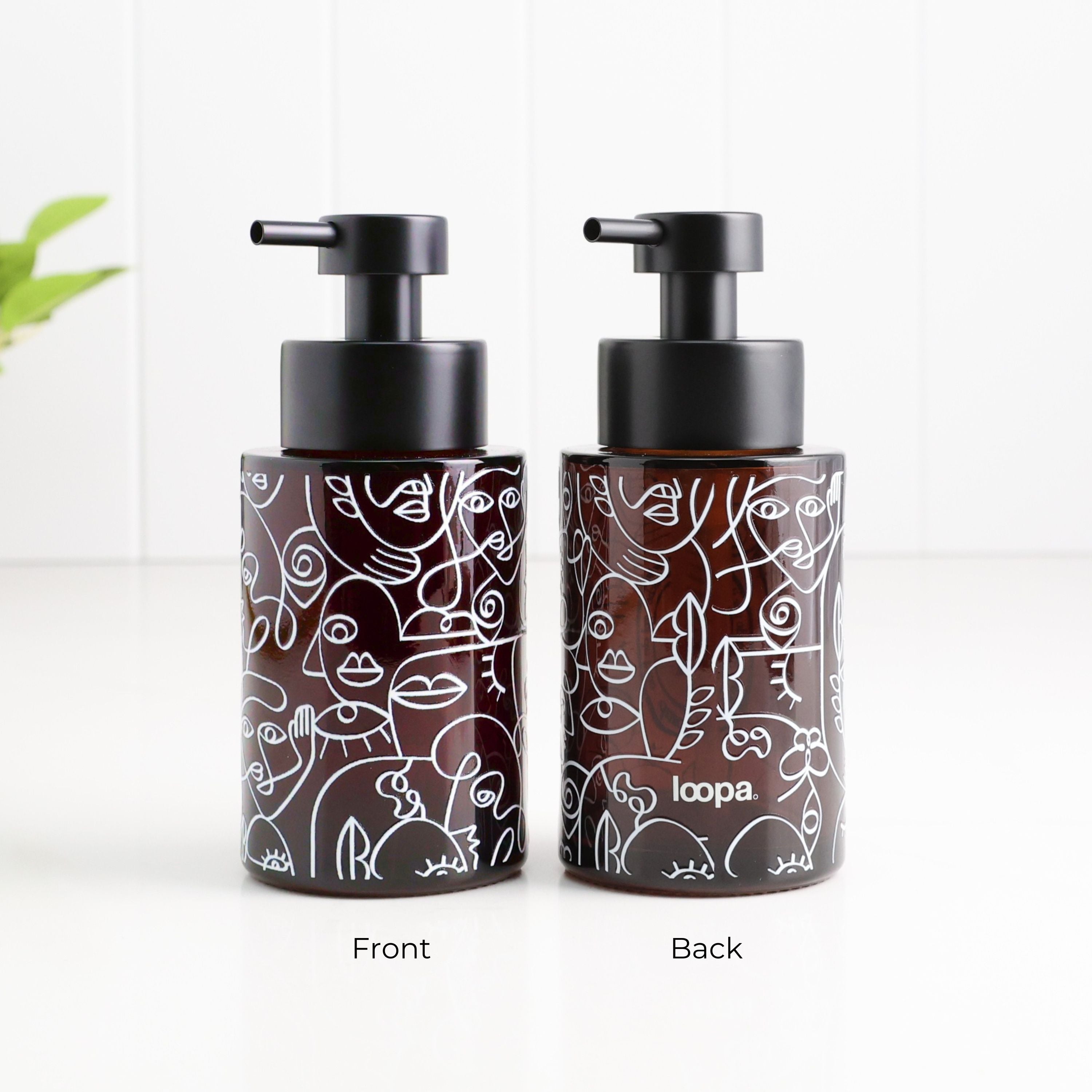 Two amber glass pump bottles with black pumps, shown side by side on a white surface against a white paneled background. The front bottle shows a white line-art pattern, and the back bottle shows the brand name 'loopa' and the reverse side of the pattern.