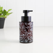 A dark brown glass pump bottle with a black pump dispenser, decorated with white minimalist line art of faces, set on a white surface with a white paneled wall background and a small green plant in the distance.