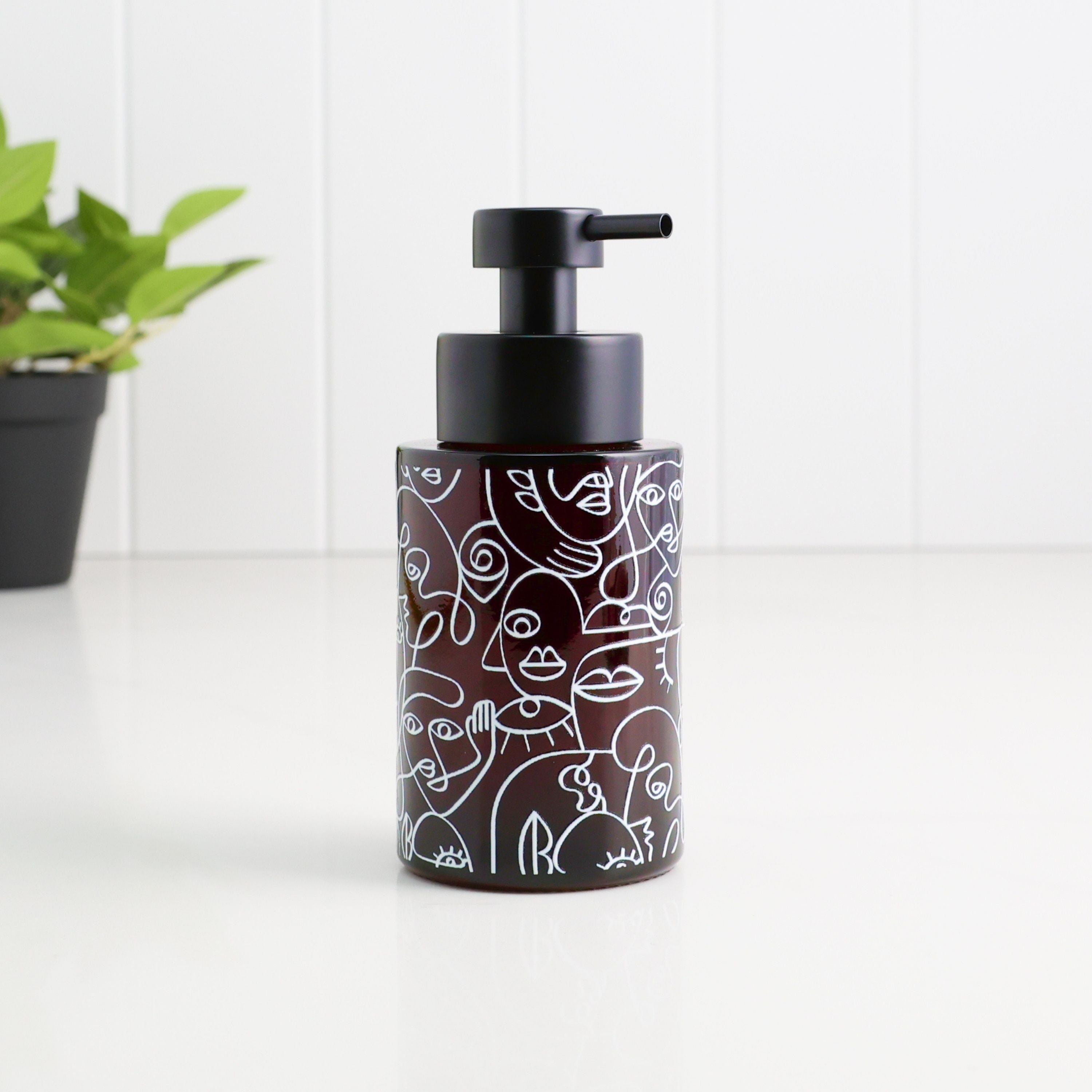 A dark brown glass pump bottle with a black pump dispenser, decorated with white minimalist line art of faces, set on a white surface with a white paneled wall background and a small green plant in the distance.