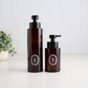 Two amber glass pump bottles with black dispensers and circular white labels, standing on a white surface against a white paneled background with a small plant at left.