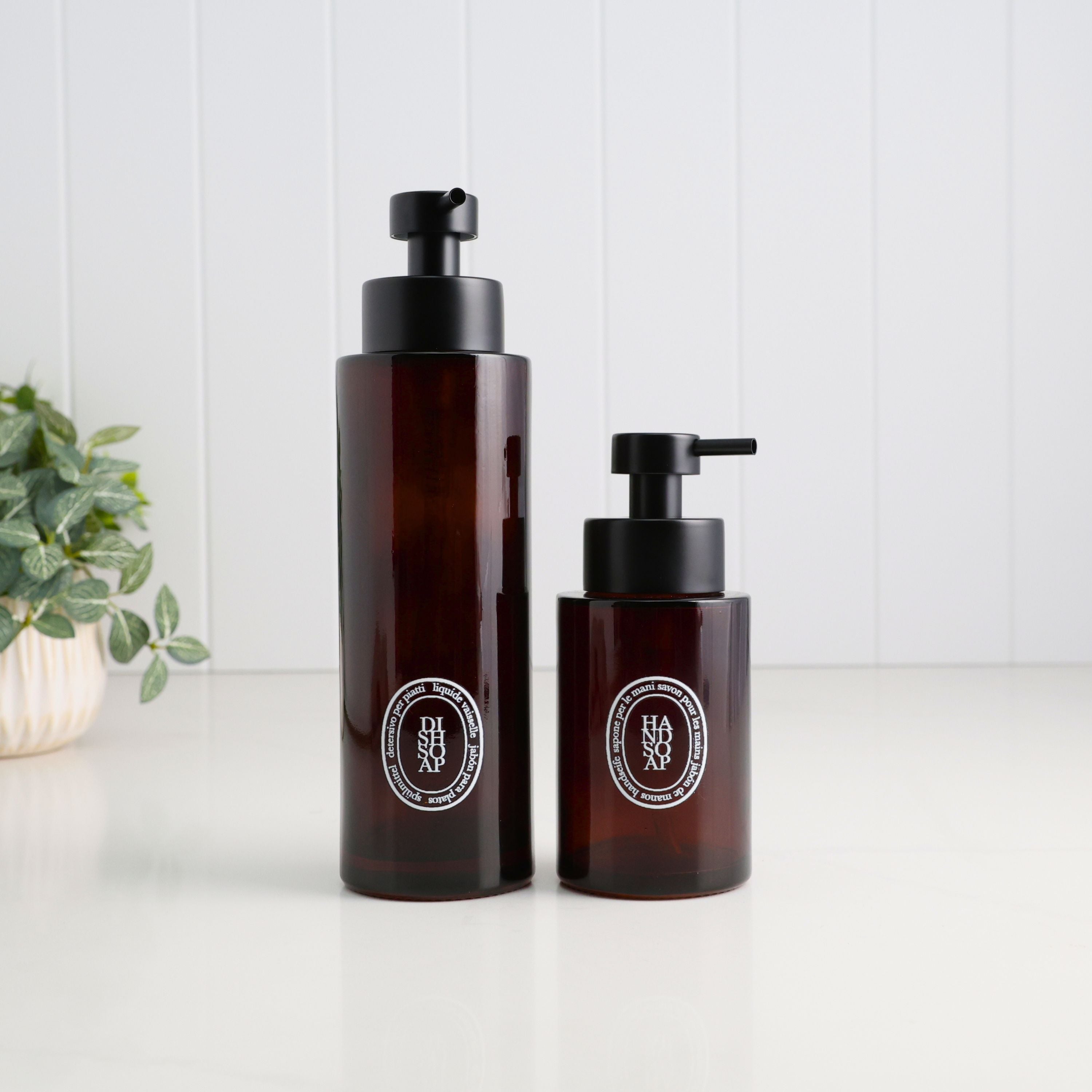 Two amber glass pump bottles with black dispensers and circular white labels, standing on a white surface against a white paneled background with a small plant at left.