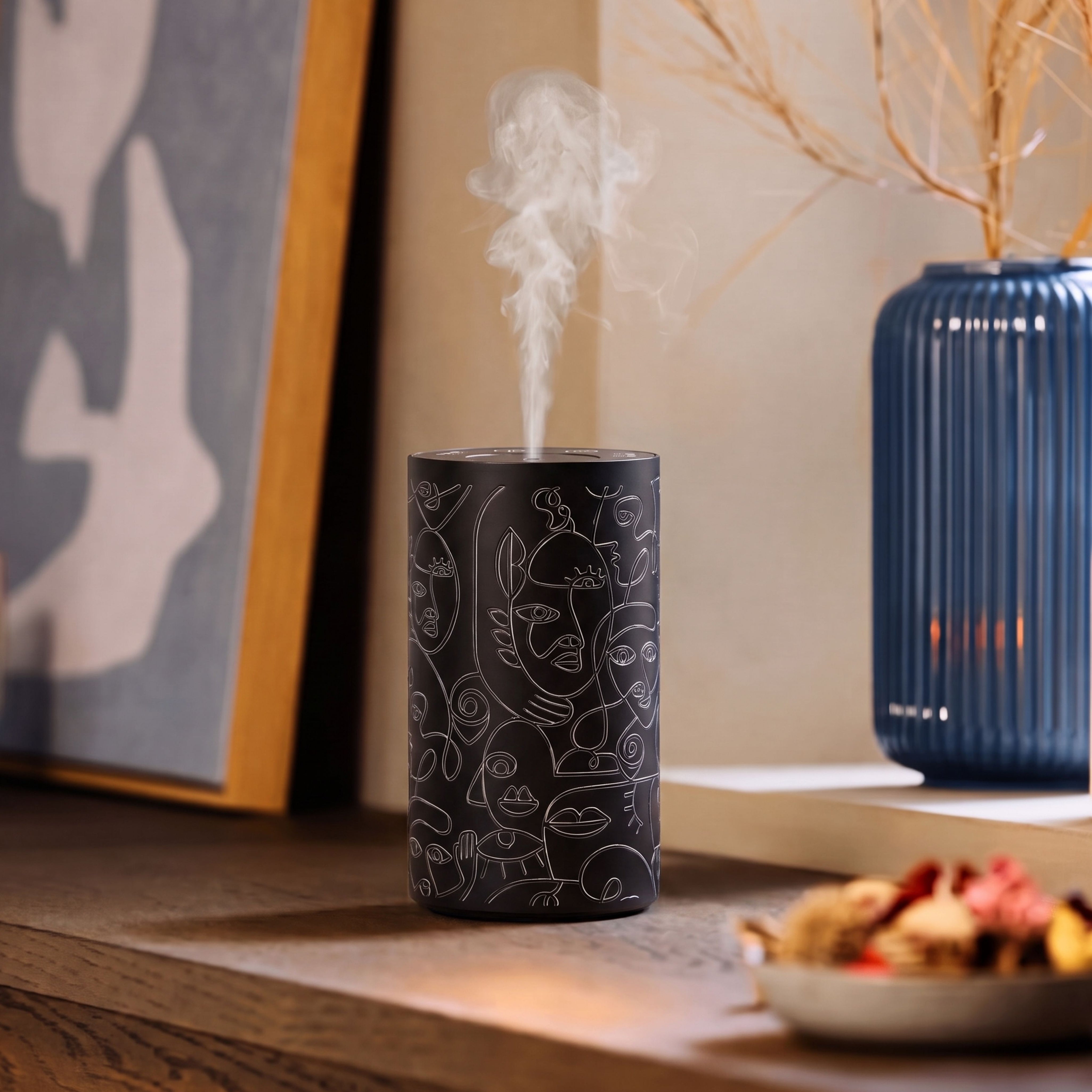 Loopa black waterless essential oil diffuser featuring abstract white line art faces, releasing a fine fragrance mist on a wooden table. Cordless, rechargeable aromatherapy diffuser with a contemporary cylindrical design, displayed beside framed artwork and a blue ceramic vase.