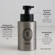 Brushed stainless steel round bottle dispenser with a black oval label and arrow detailing, designed for use in bathrooms, kitchens, or shower areas