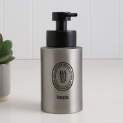 Brushed stainless steel round bottle dispenser with a black oval label, shown as a 250ml refillable bottle suited to cloakrooms, WC areas, or compact bathrooms